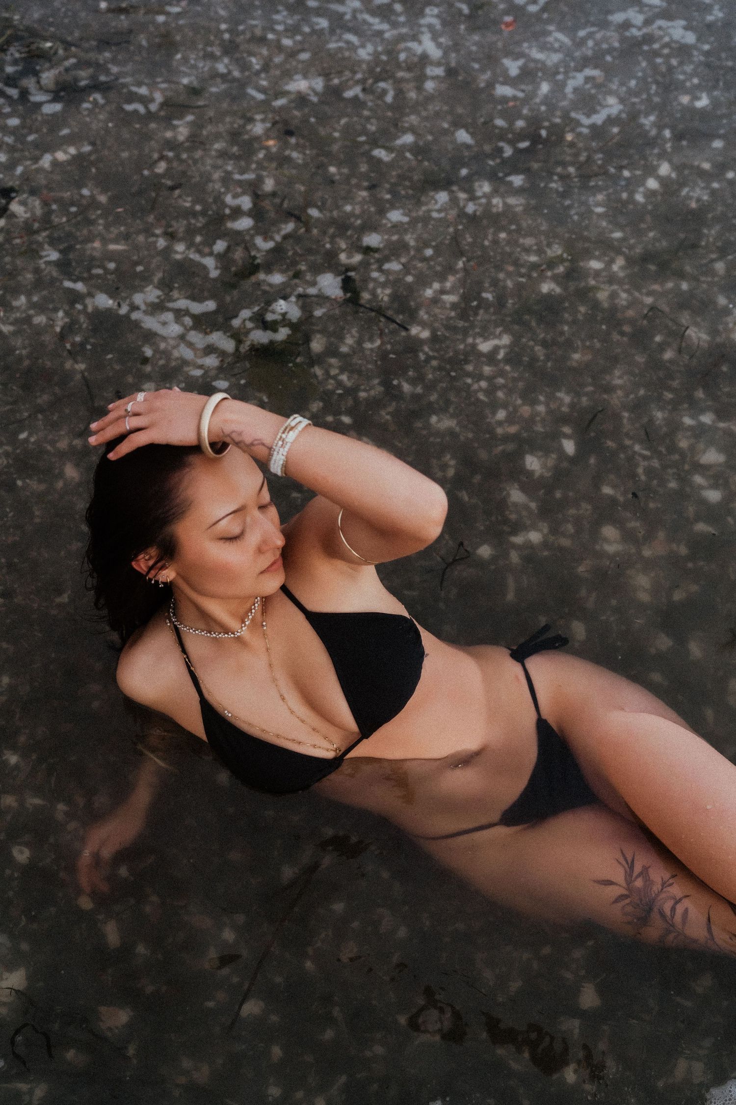 Woman in black bikini lying on wet sand at beach, wearing jewelry and showing tattoo on leg.