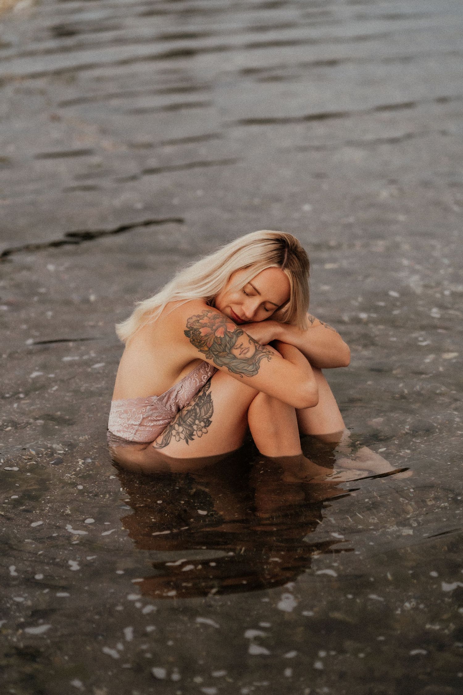 Tattooed blonde woman sitting in shallow water with eyes closed, hugging her knees in a serene outdoor setting.