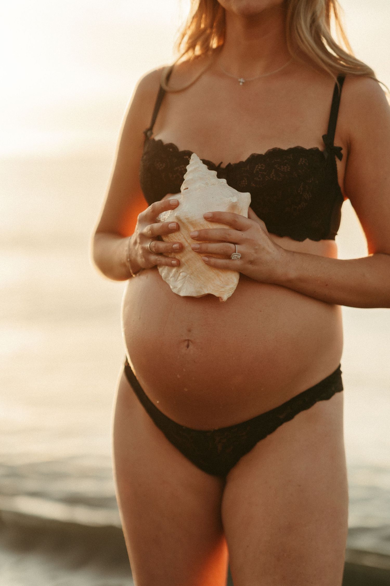 Pregnant woman in black lace lingerie holding a white seashell on a beach at golden hour sunset.