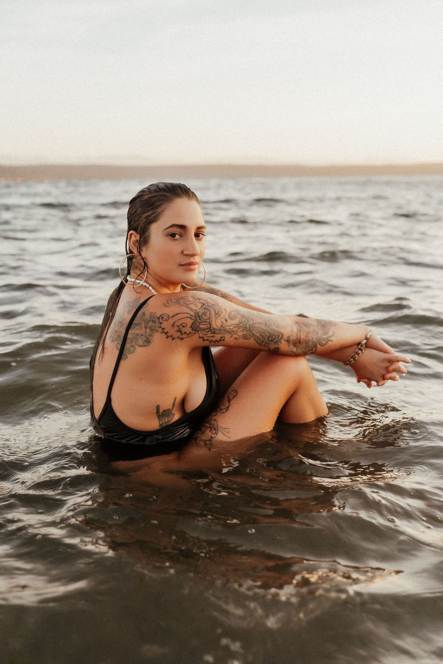 Tattooed woman in black swimsuit sitting in shallow ocean water at sunset, looking over shoulder.