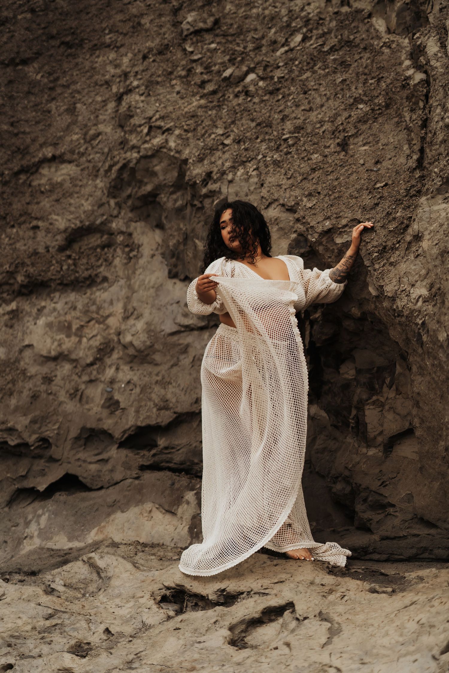Figure in flowing white lace dress poses gracefully against dramatic rocky cliff backdrop.