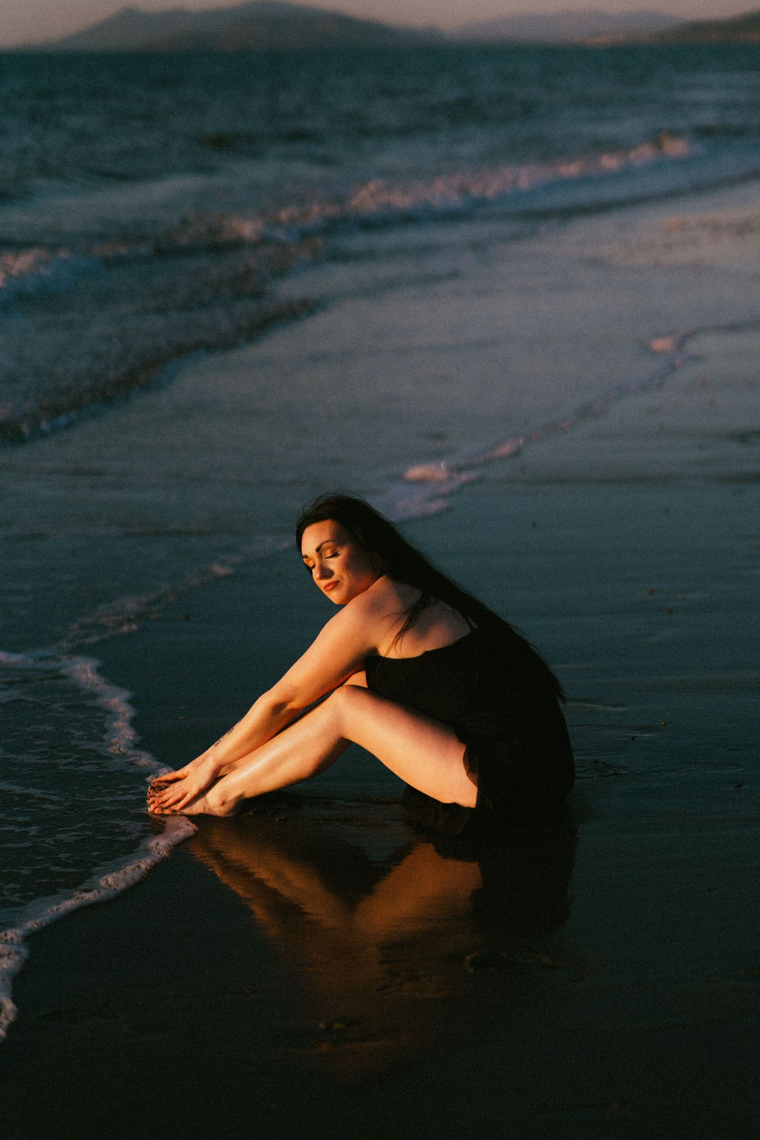 A person in dark clothing sits reflectively on wet sand at the beach during sunset, creating a moody silhouette.