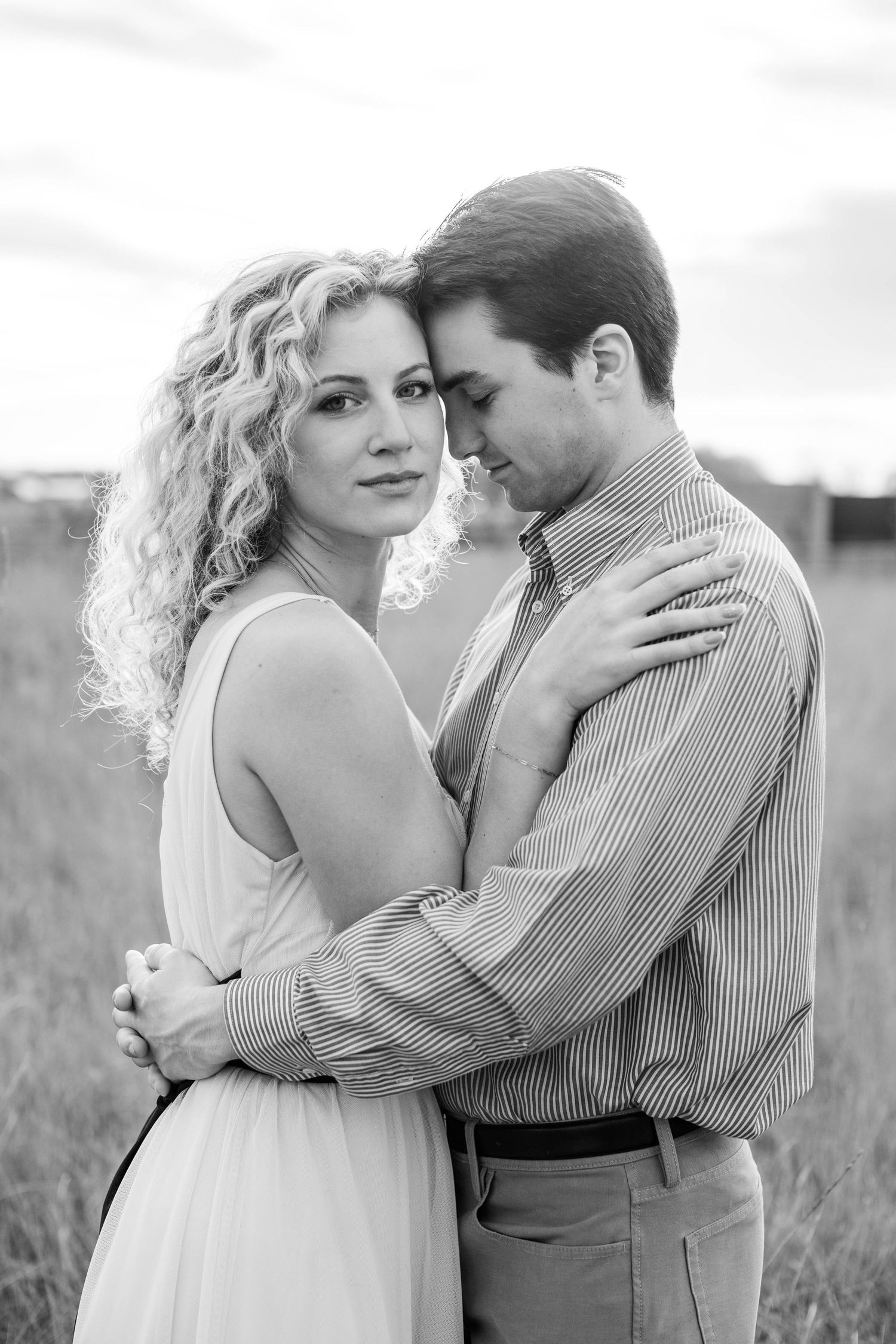 Black and white photo of a romantic couple embracing in a field, woman in white dress with curly hair looking at camera.