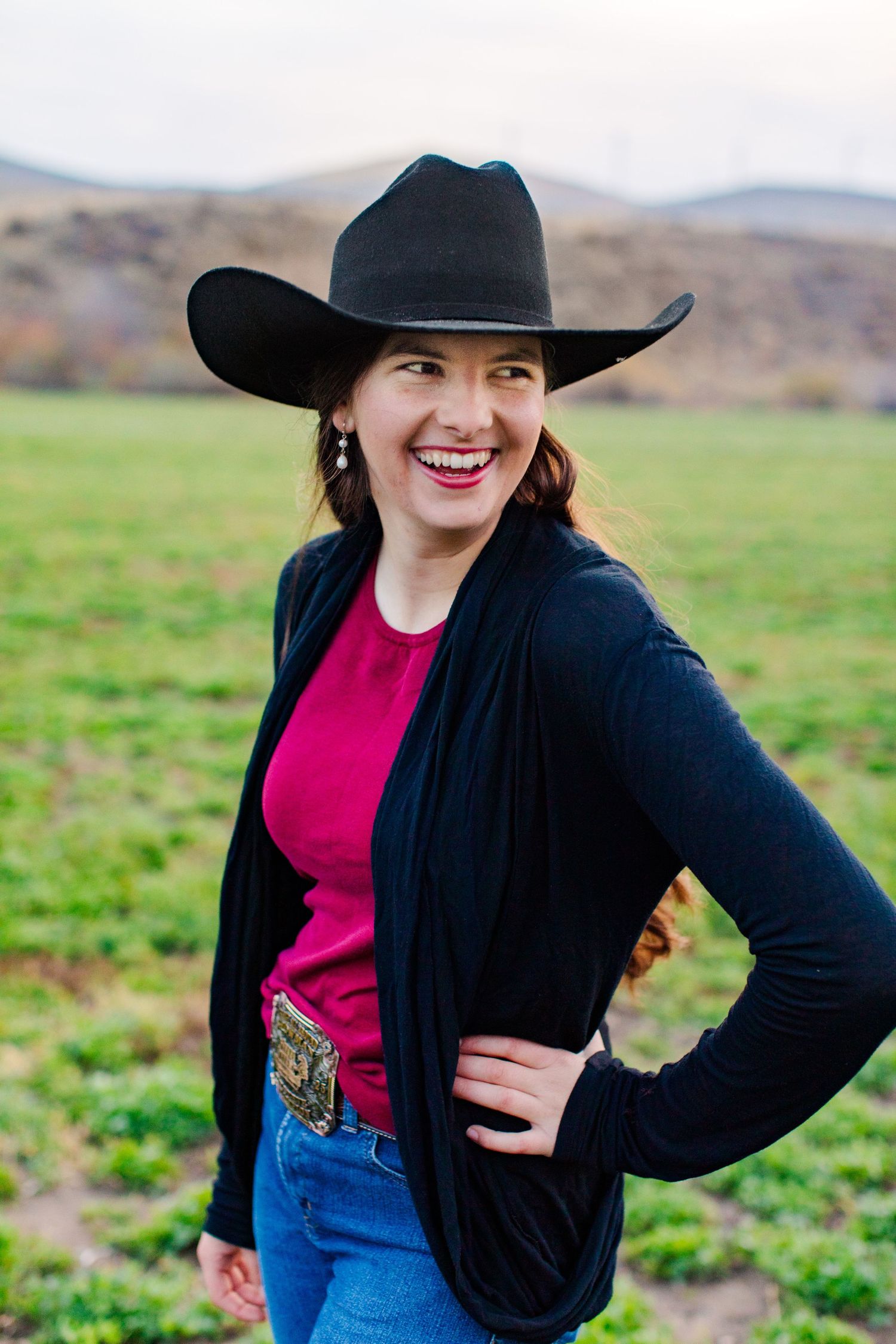 Woman in western attire with black hat and cardigan poses outdoors against rolling hills.