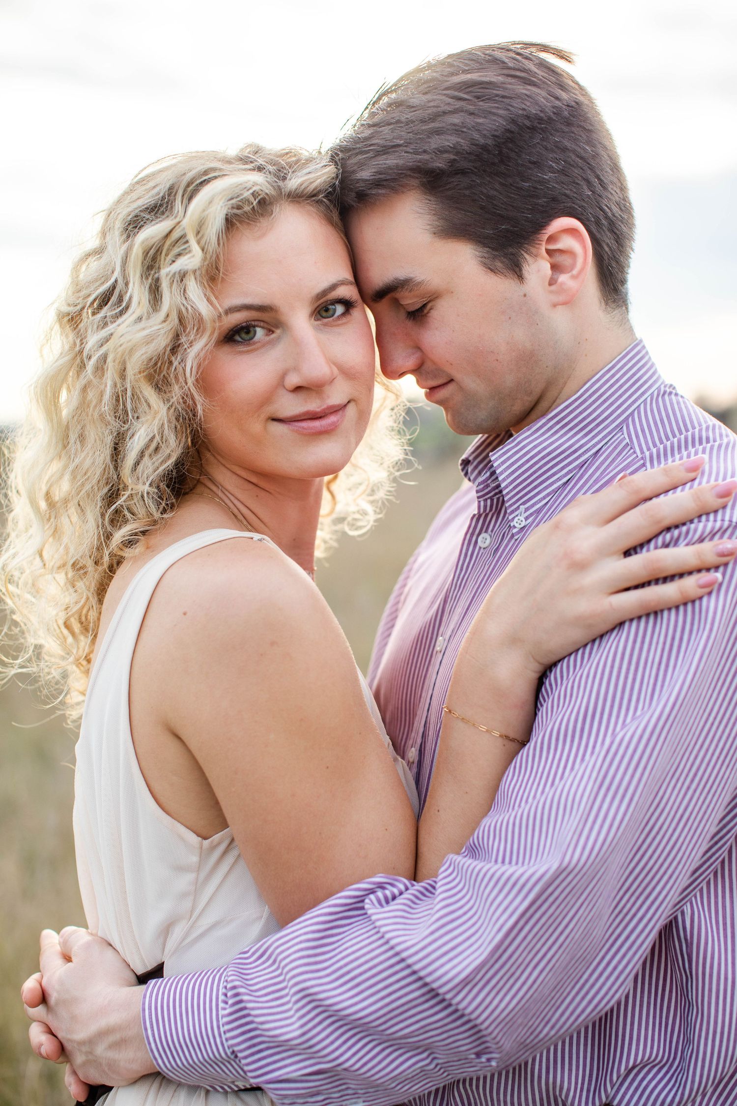 Romantic couple embracing outdoors, blonde curly-haired woman in white dress and man in purple striped shirt.