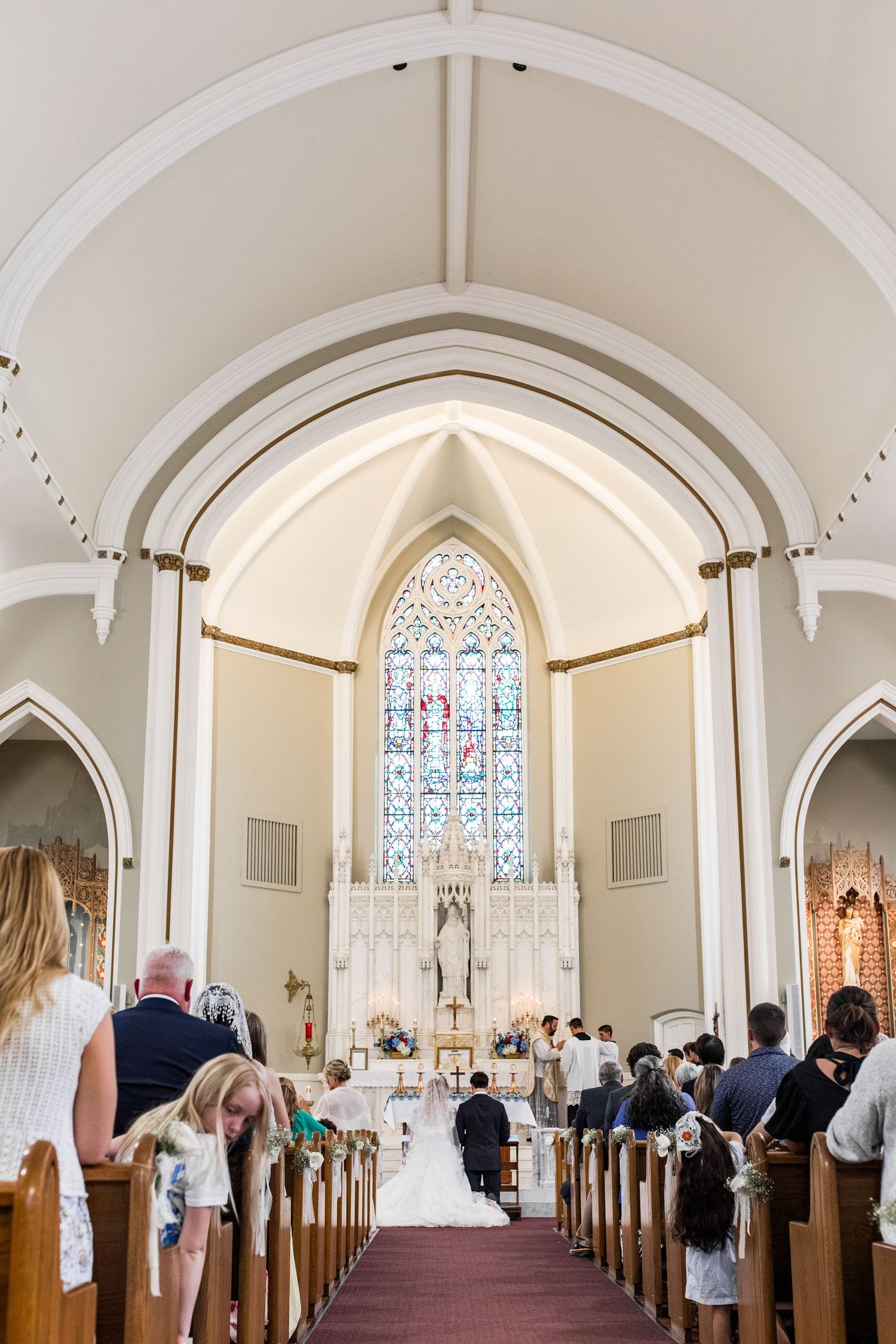 A wedding ceremony inside a white Gothic church with stained glass windows, guests seated in pews, and couple at the altar.