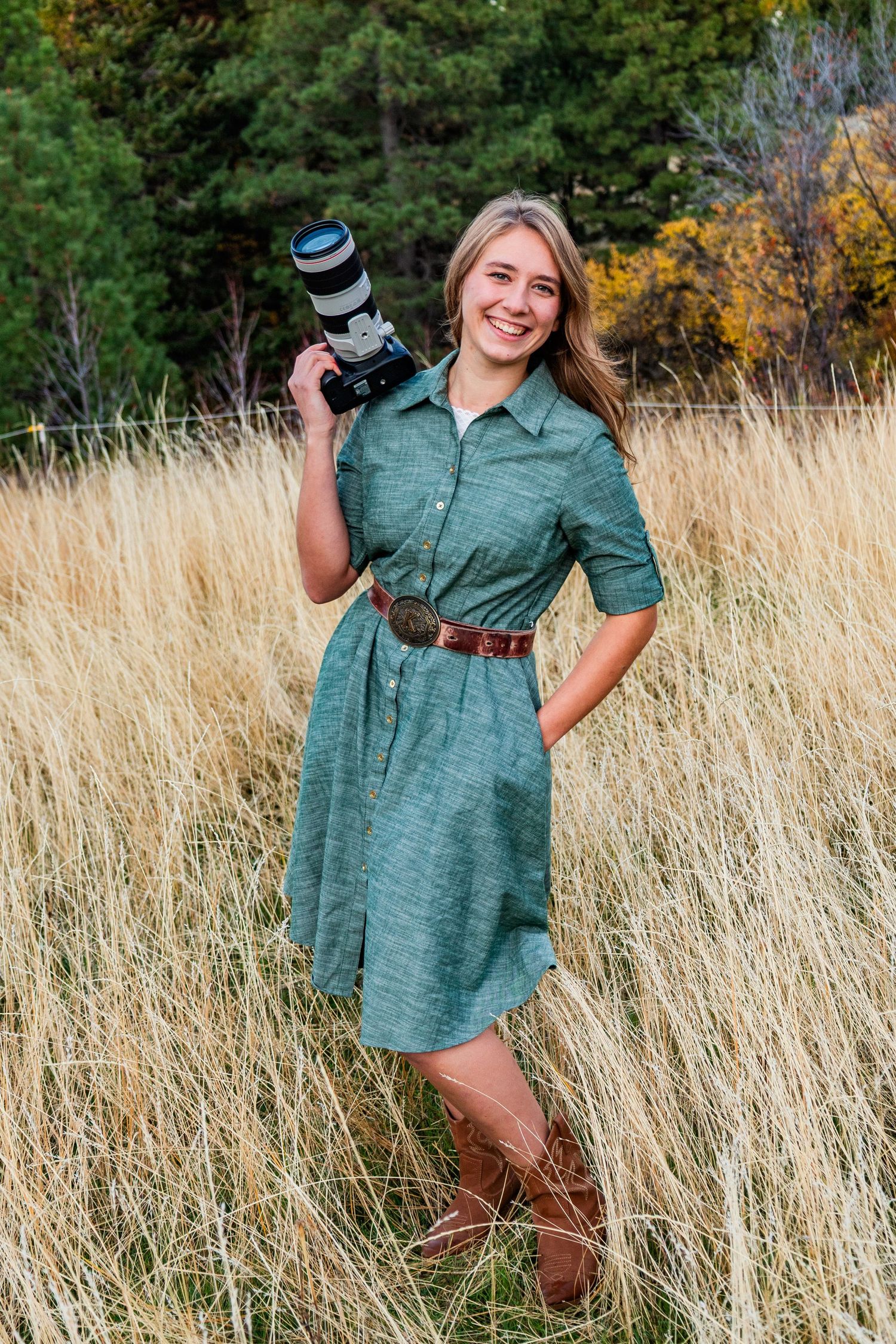 A green dress flows in the breeze among tall golden grass with autumn trees in the background.