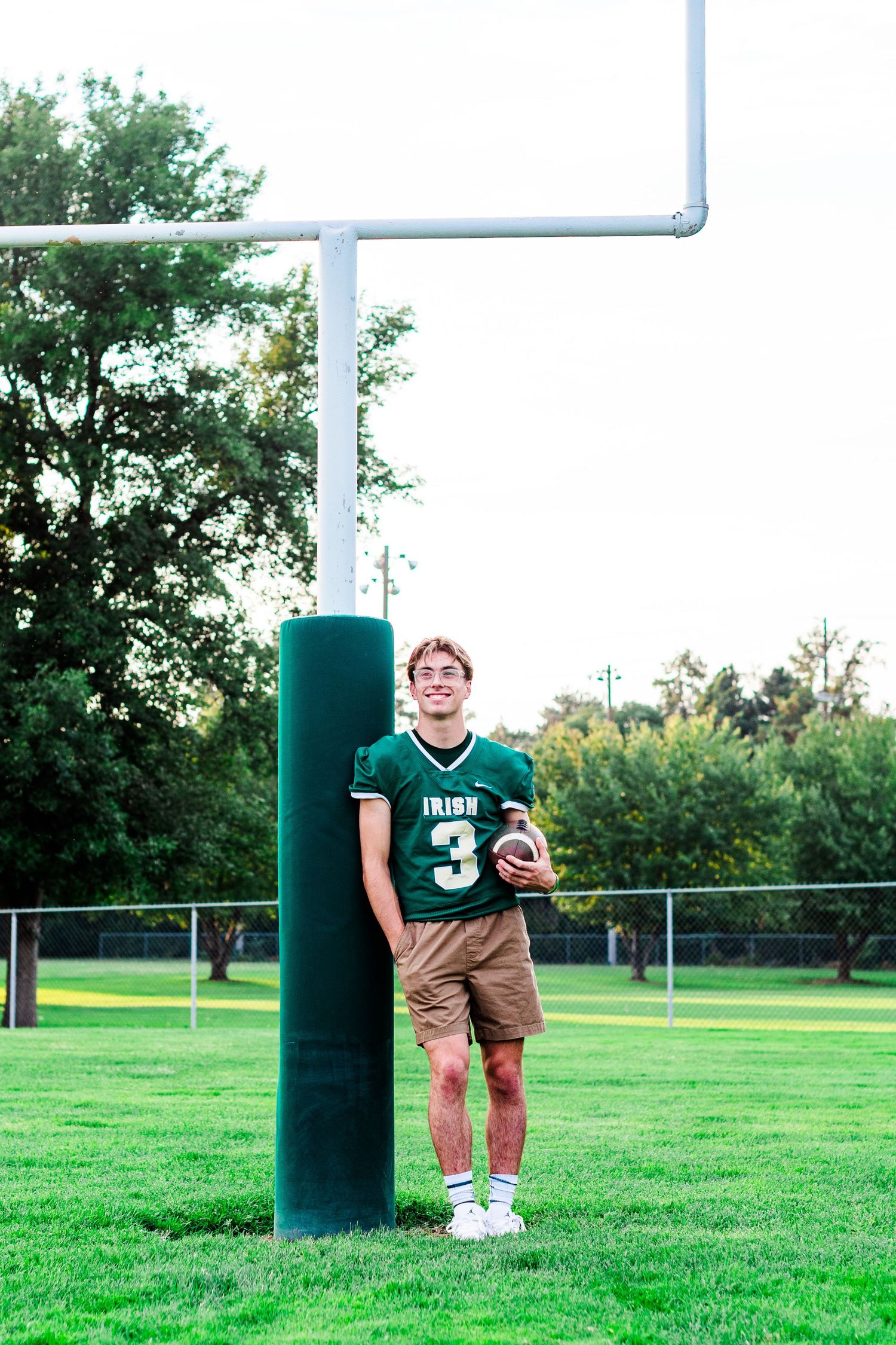 Football player in green Irish jersey and khaki shorts poses with ball on grassy field near goal post.