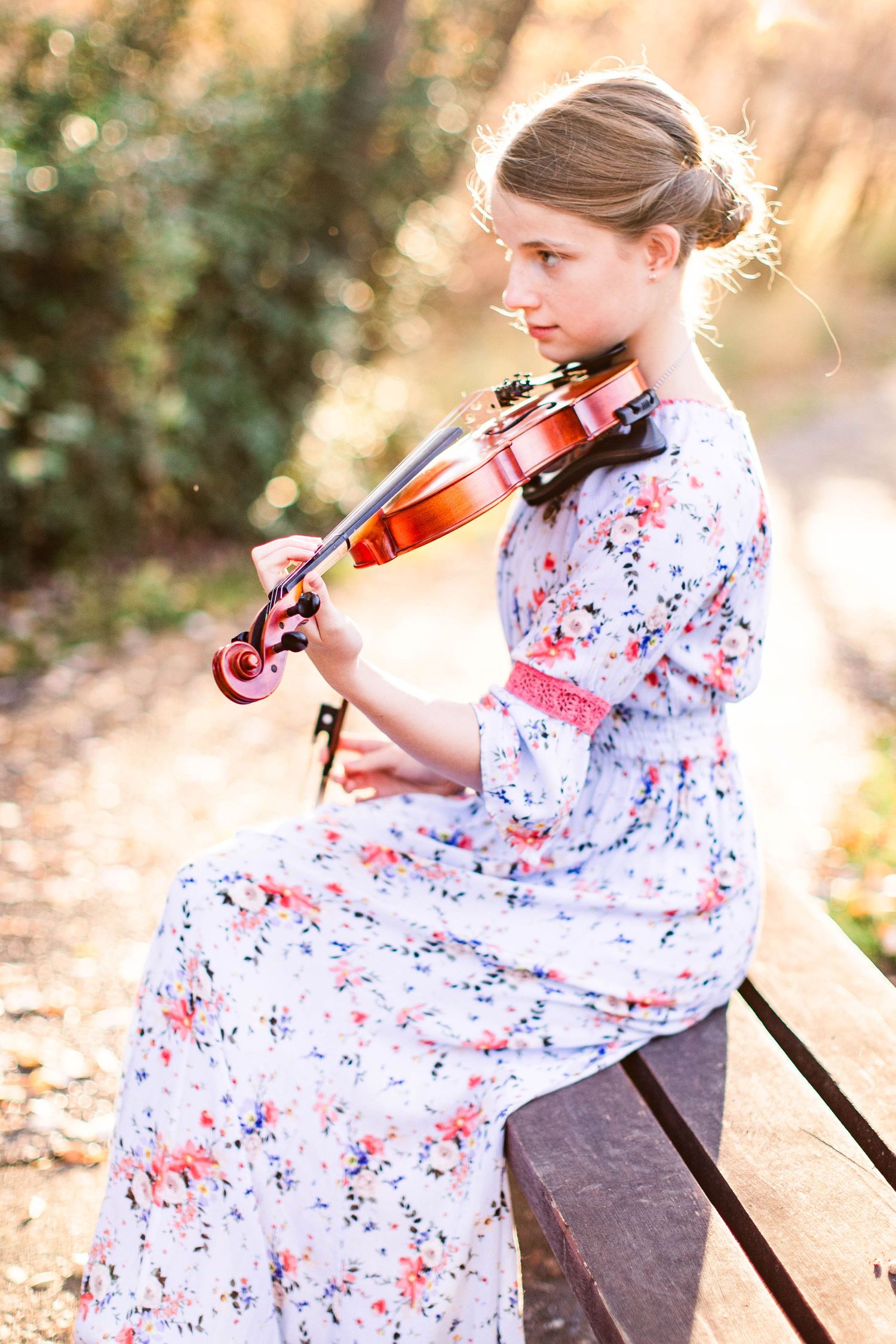 A violinist in a floral dress performing on a wooden bench in natural lighting.