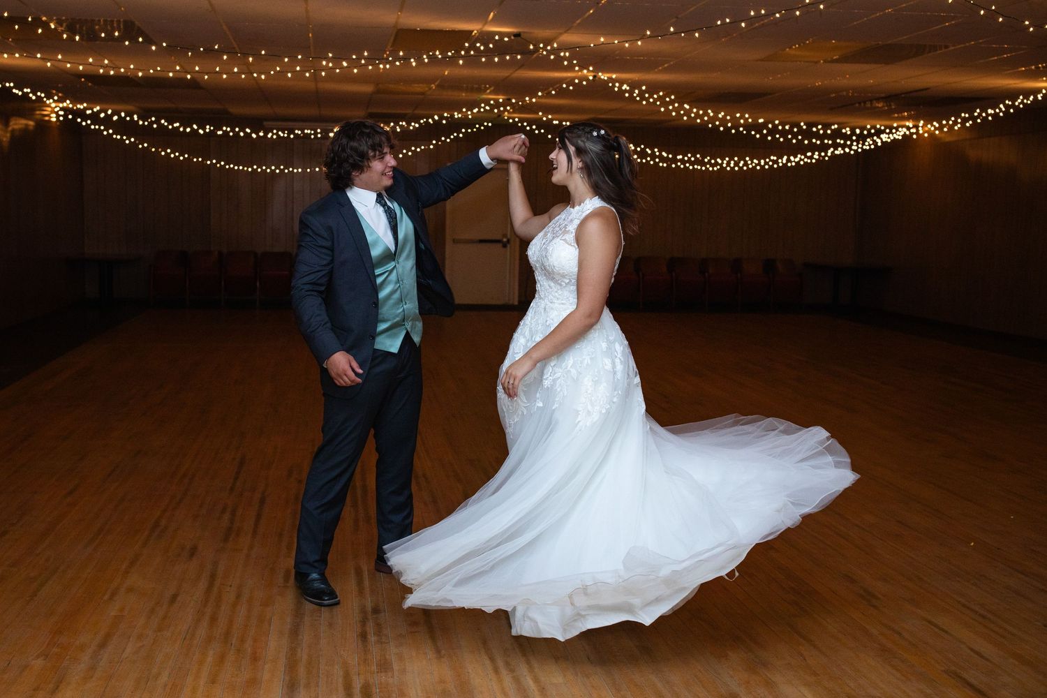 Wedding couple dances under twinkling string lights on a wooden dance floor.