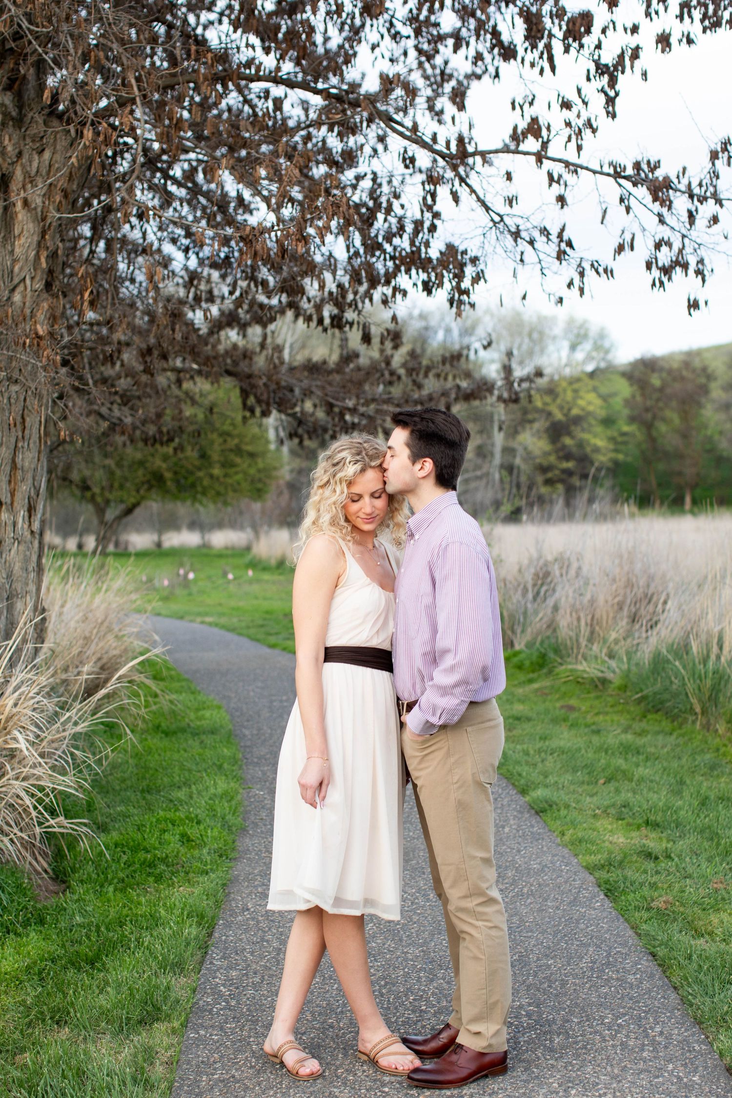 A man in a lavender shirt kisses a woman in a white dress on a garden pathway surrounded by lush greenery.