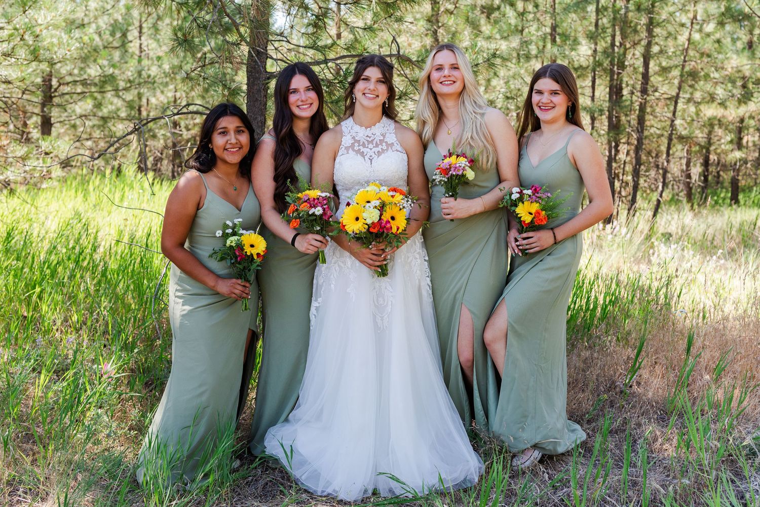 Bridal party in sage green dresses poses together in a forest setting.