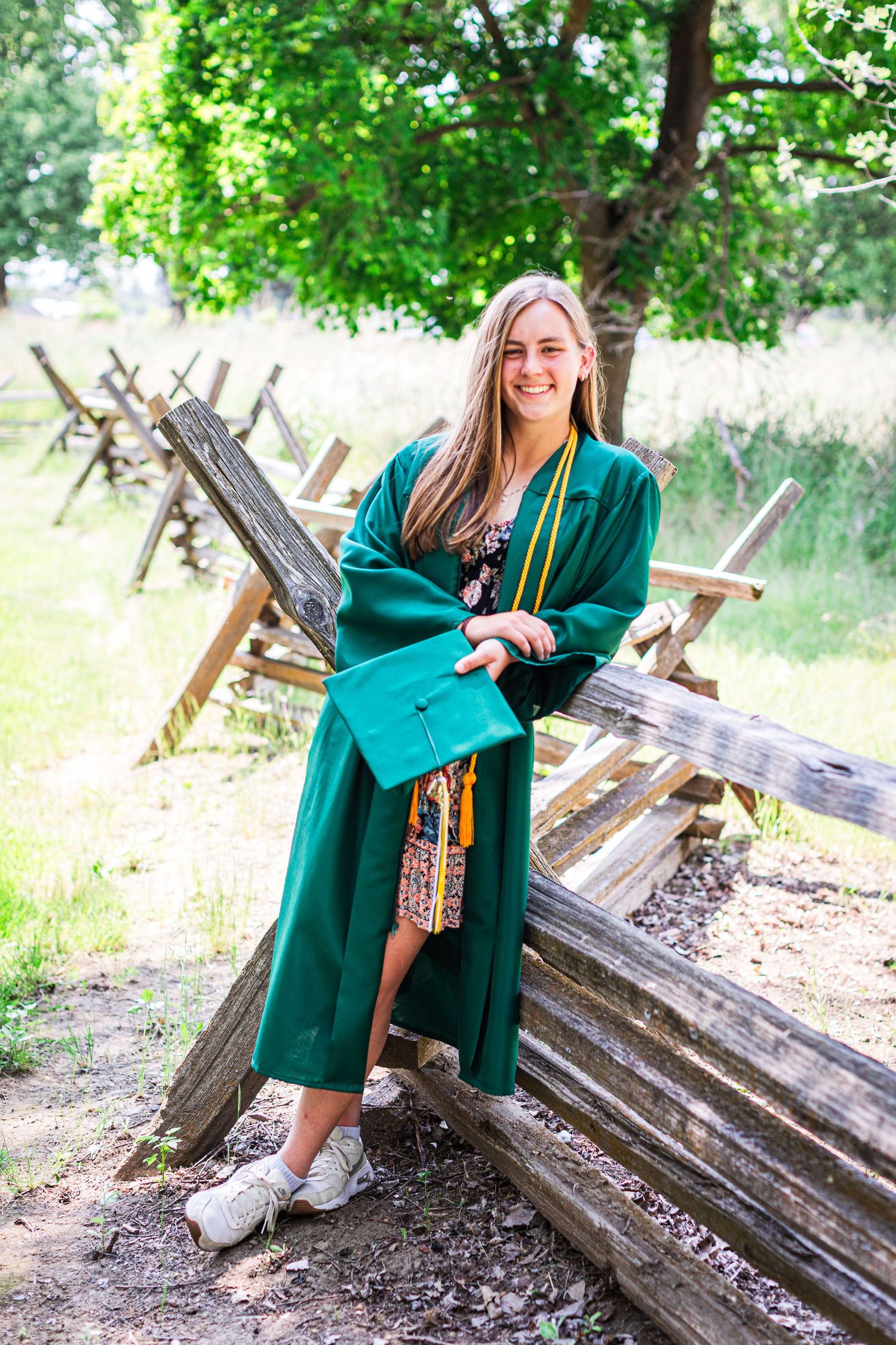 A DeSales Catholic High School graduate in a green dress sits on rustic wooden logs in a sunny outdoor setting.