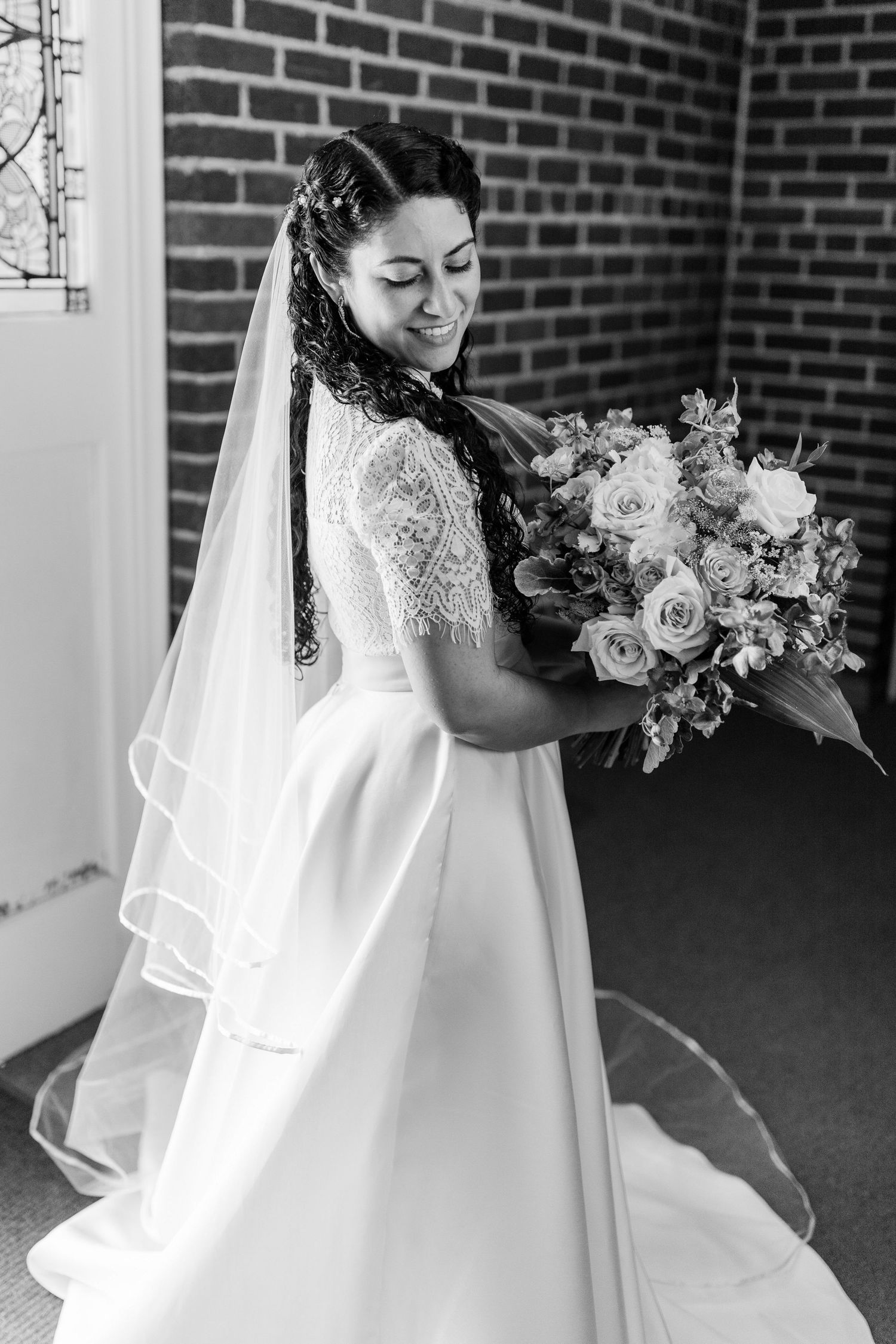 A bride in a white lace wedding dress and veil holds a romantic bouquet of roses against a brick wall backdrop.