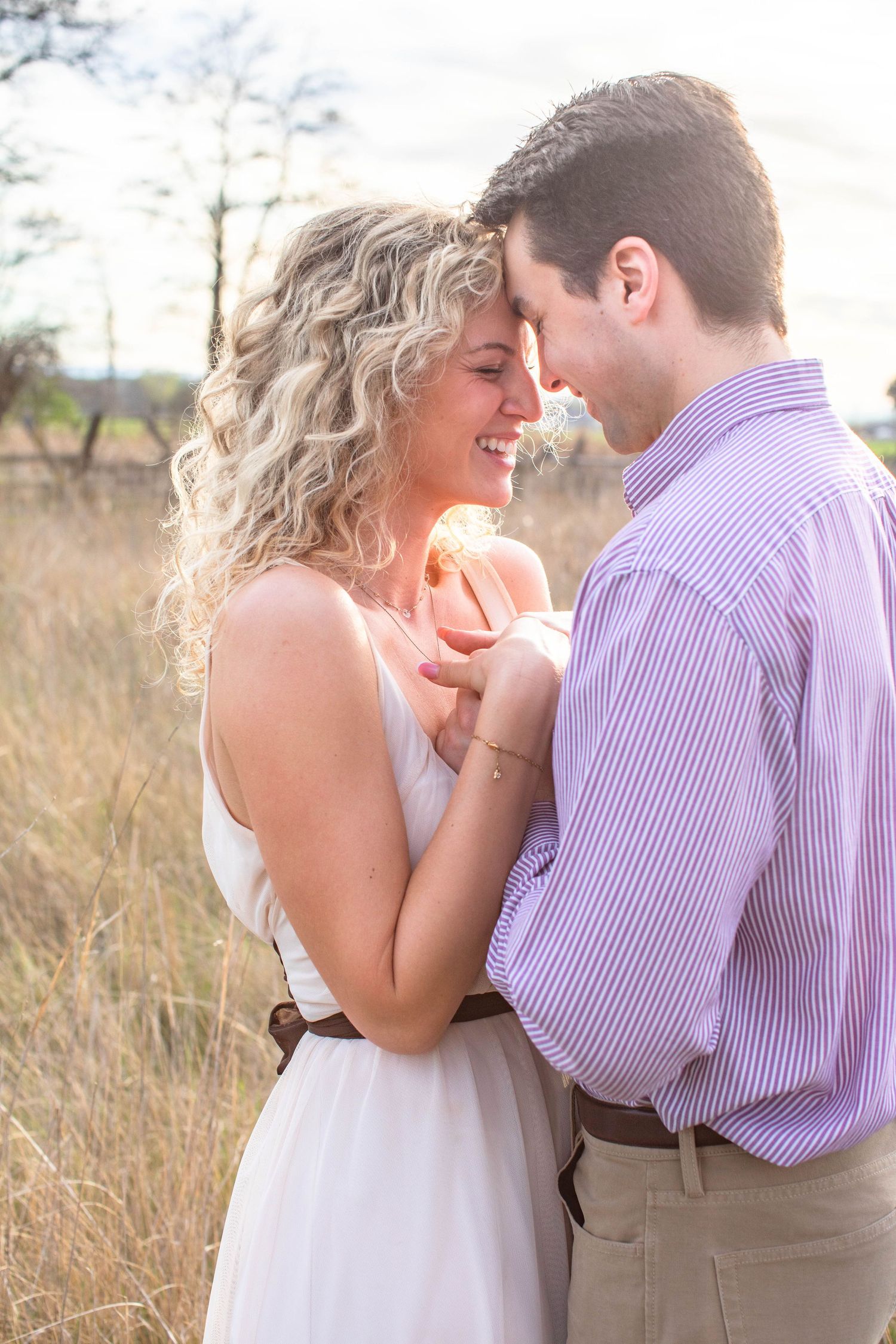 Couple embracing in golden field at sunset, woman in white dress laughing as man leans forehead to hers.