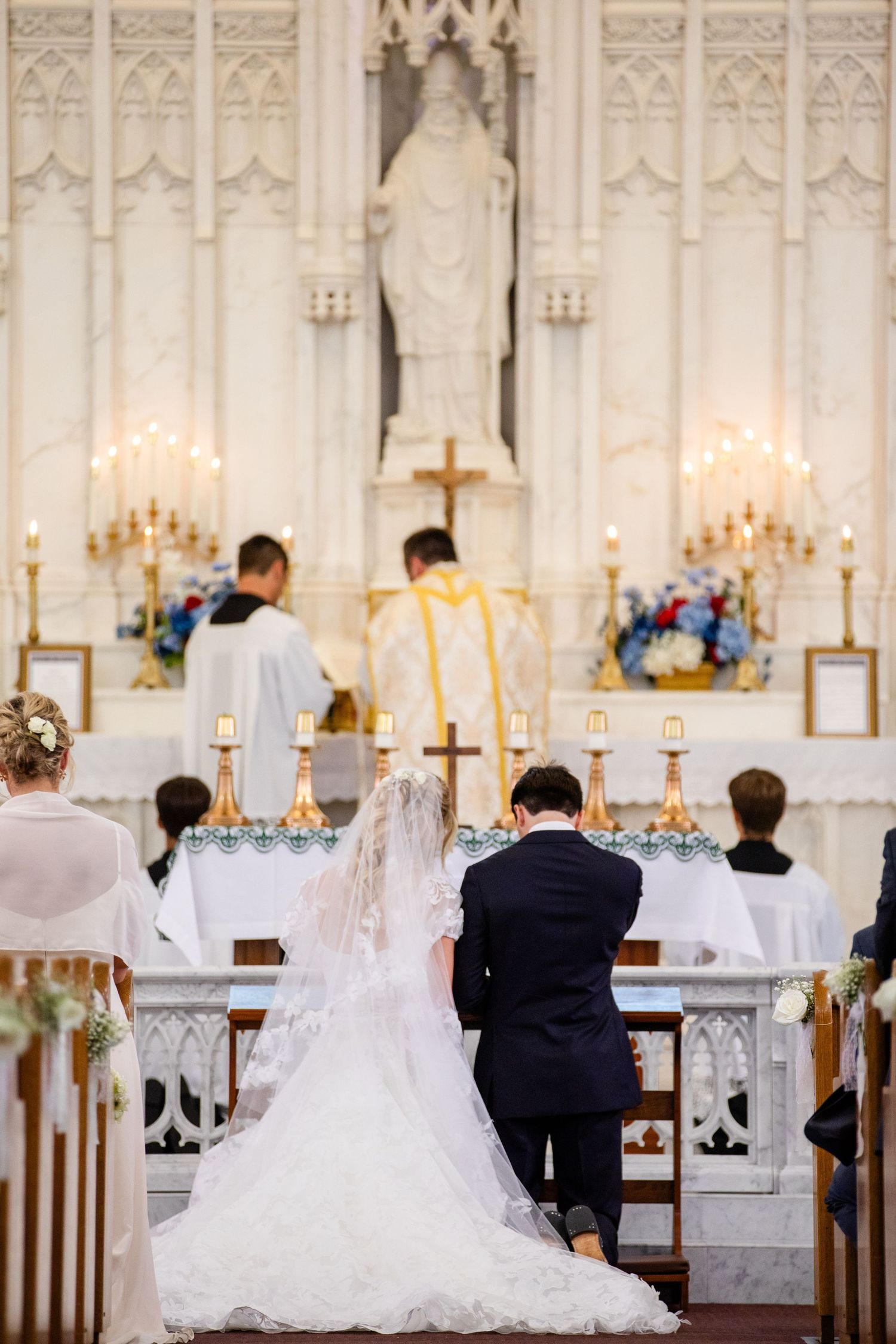 Bride and groom kneeling at church altar during Catholic wedding ceremony with priest officiating.