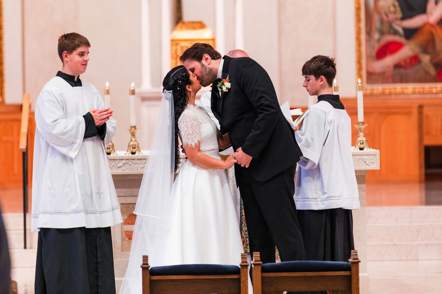 A couple shares their first kiss during a Catholic wedding ceremony with altar servers in white robes standing beside them.
