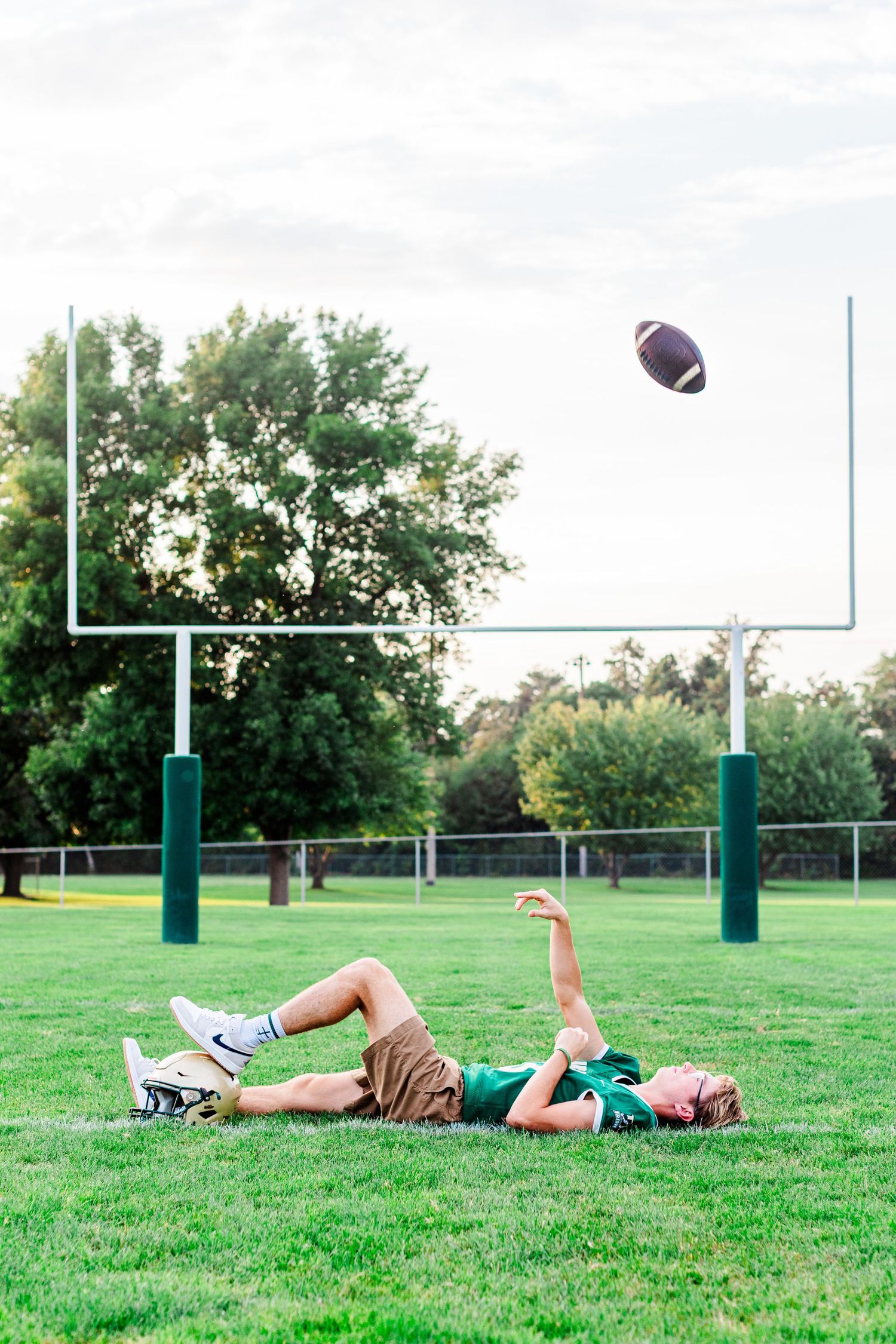 Player laying on grass field throwing football towards goal post during practice.