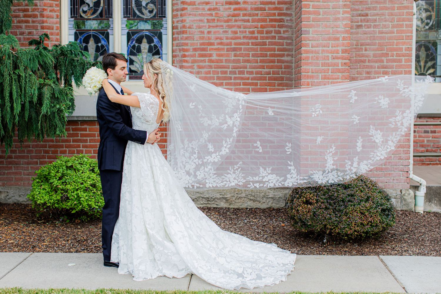 Bride and groom share a romantic kiss outdoors, with her lace veil flowing in the wind against a brick church.