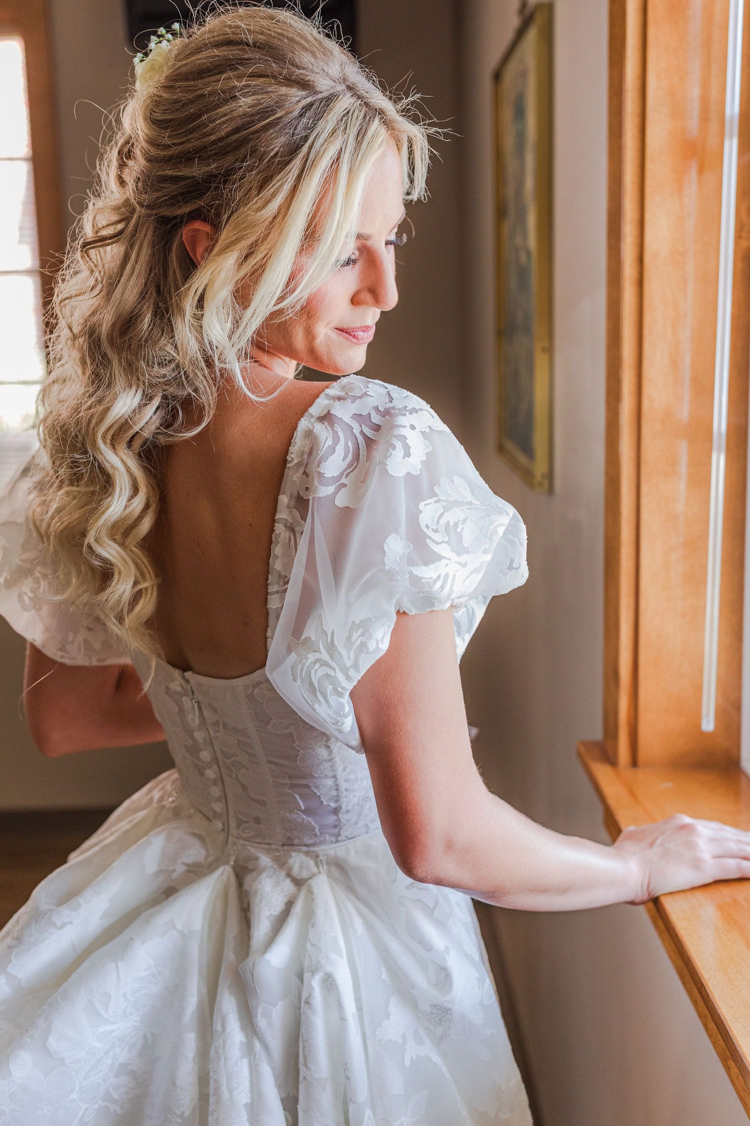 Bride in elegant white lace wedding gown with puff sleeves and open back, gazing out a window with curled blonde hair.