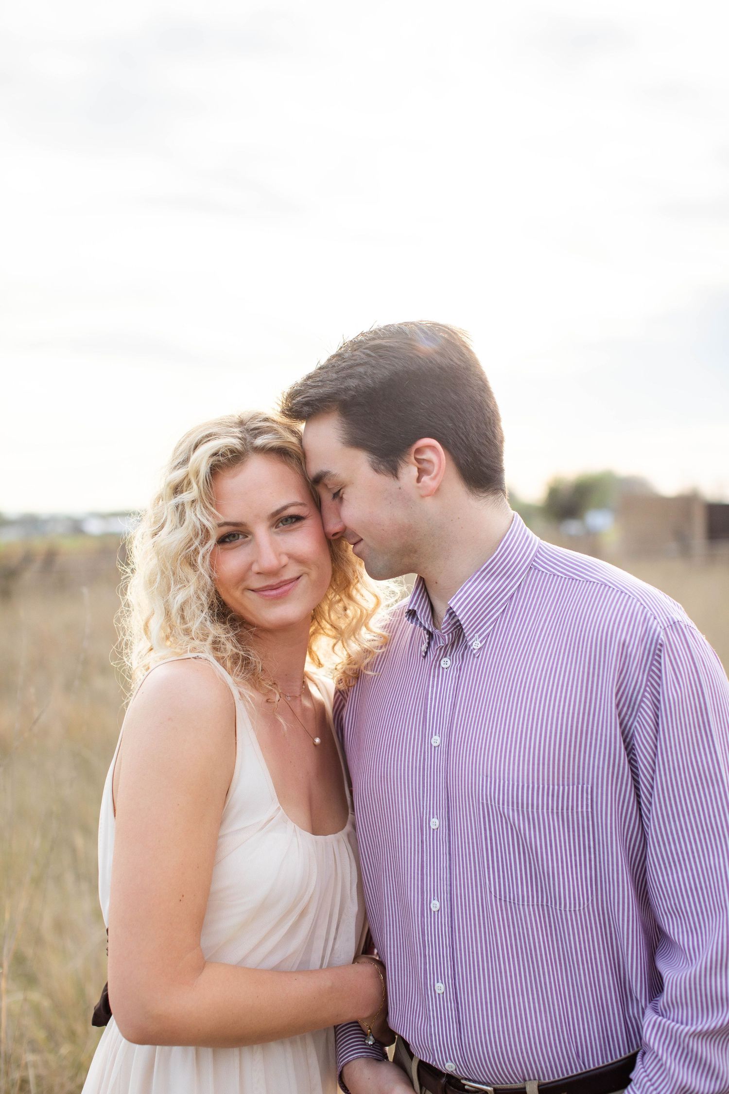 A romantic couple embraces outdoors in a golden field, the blonde woman in white dress smiling warmly as her partner leans close.