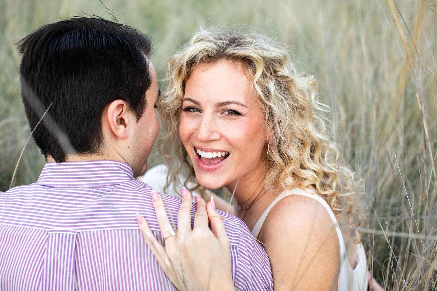 A joyful blonde woman laughs with a man in a striped shirt outdoors, showcasing an engagement ring.