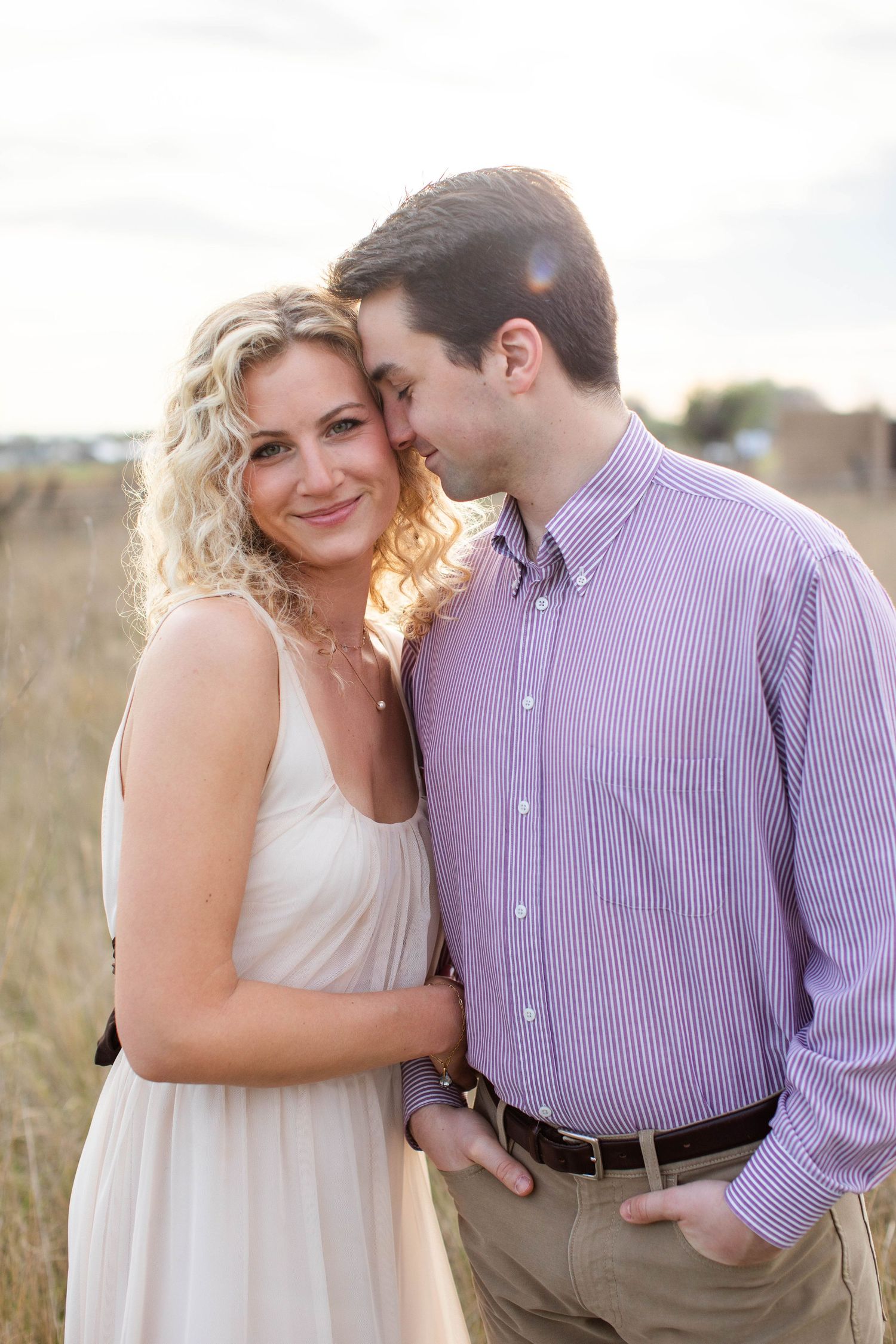 A couple embraces in a golden field, woman in white dress smiling, man in purple shirt nuzzling her cheek.