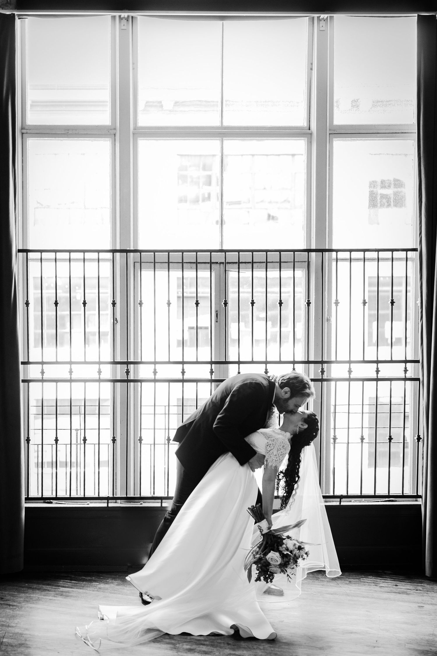 Dramatic black and white wedding portrait of couple in romantic dip pose by window.