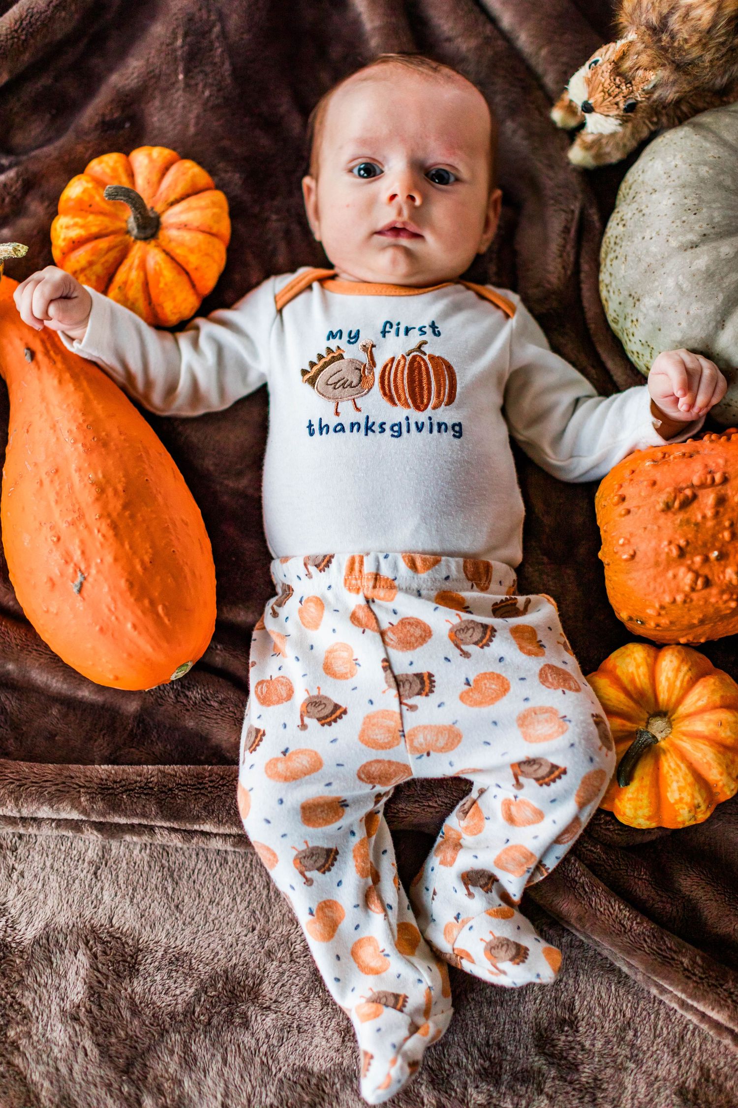 Infant wearing white and orange pumpkin-themed Thanksgiving outfit lying among decorative pumpkins on brown fabric.