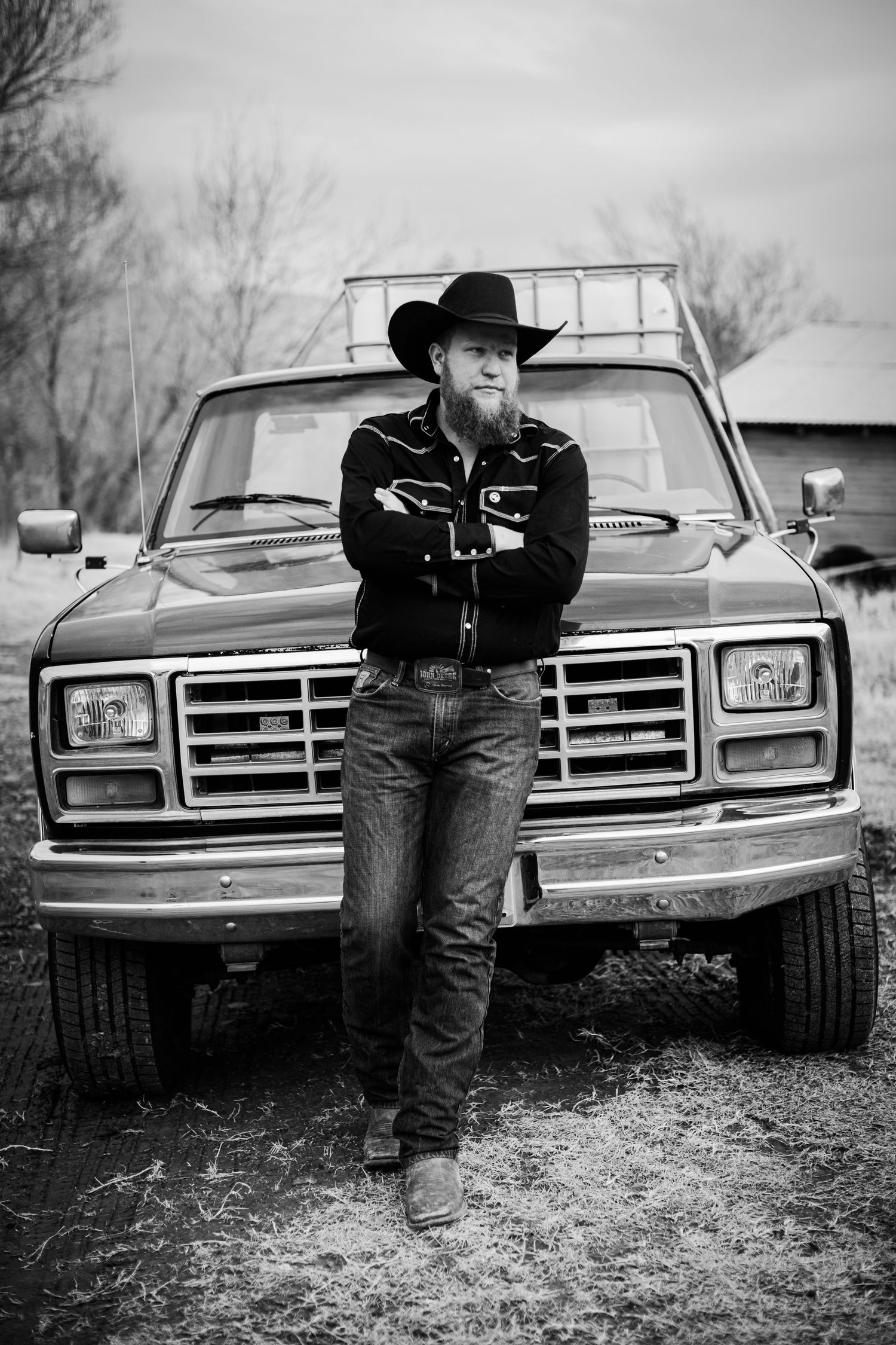 Person in western wear leans against vintage pickup truck in black and white photo.