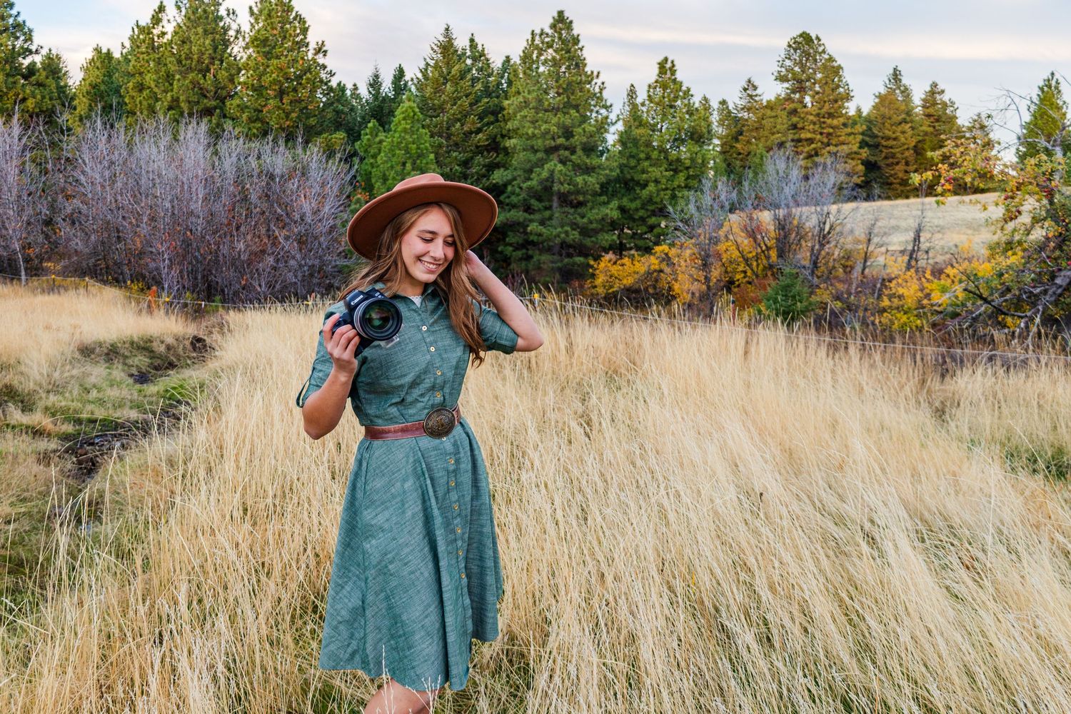 A teal dress stands out against golden autumn grass and evergreen trees in a scenic outdoor photoshoot.