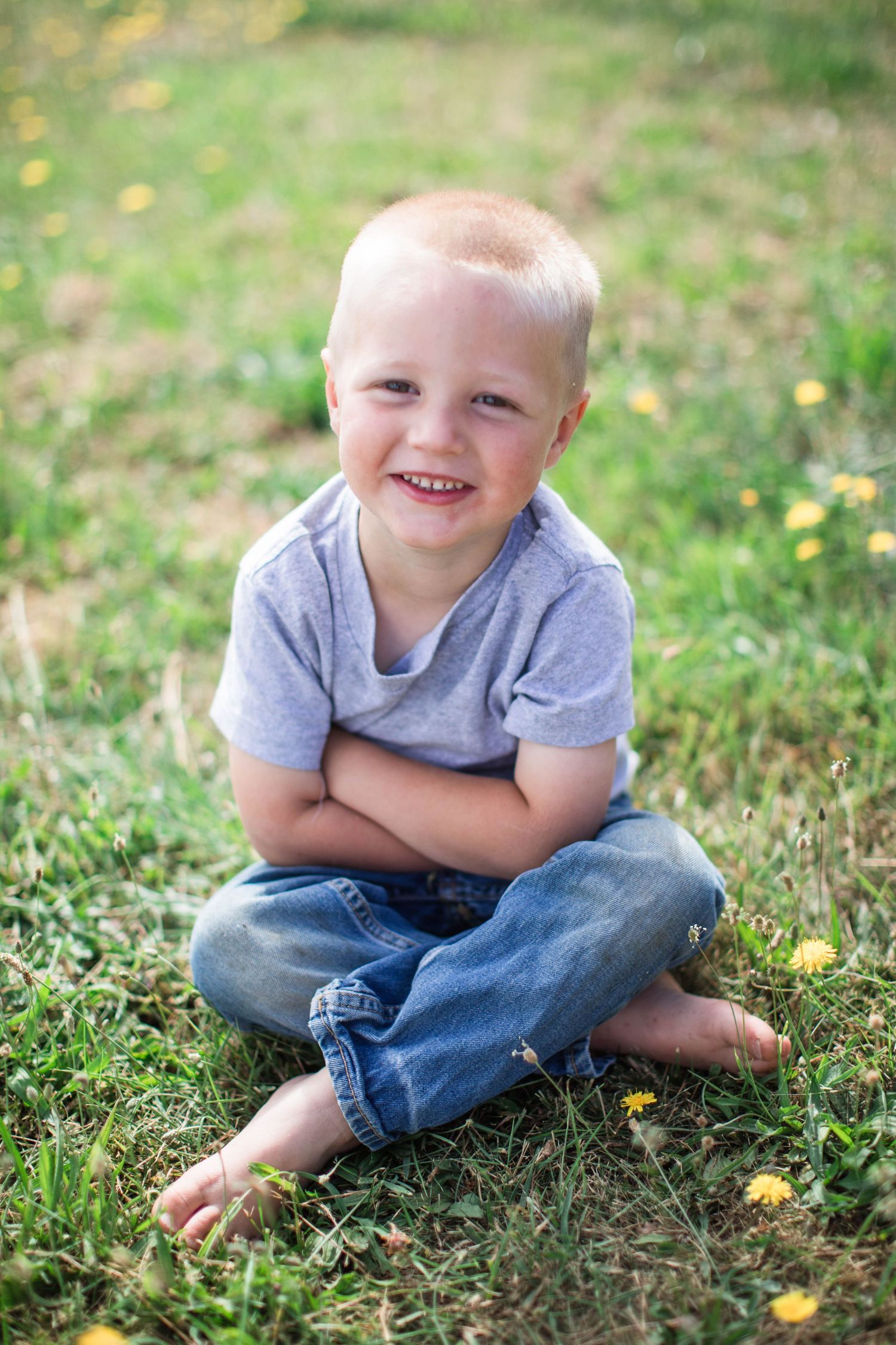 Young child sitting cross-legged on grass with dandelions wearing jeans and grey shirt.
