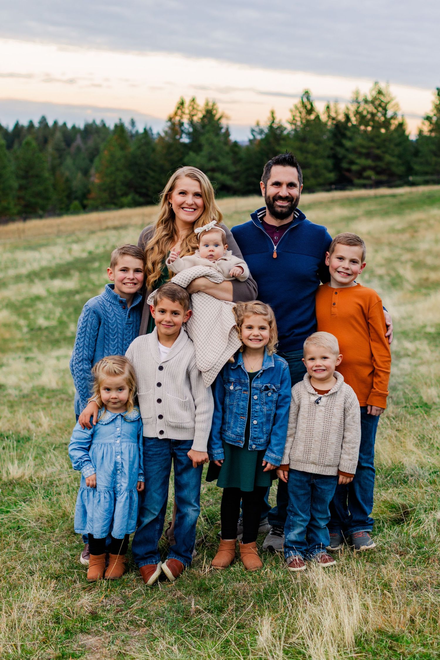 A family wearing coordinated blue and neutral toned outfits poses together in a grassy meadow at sunset.