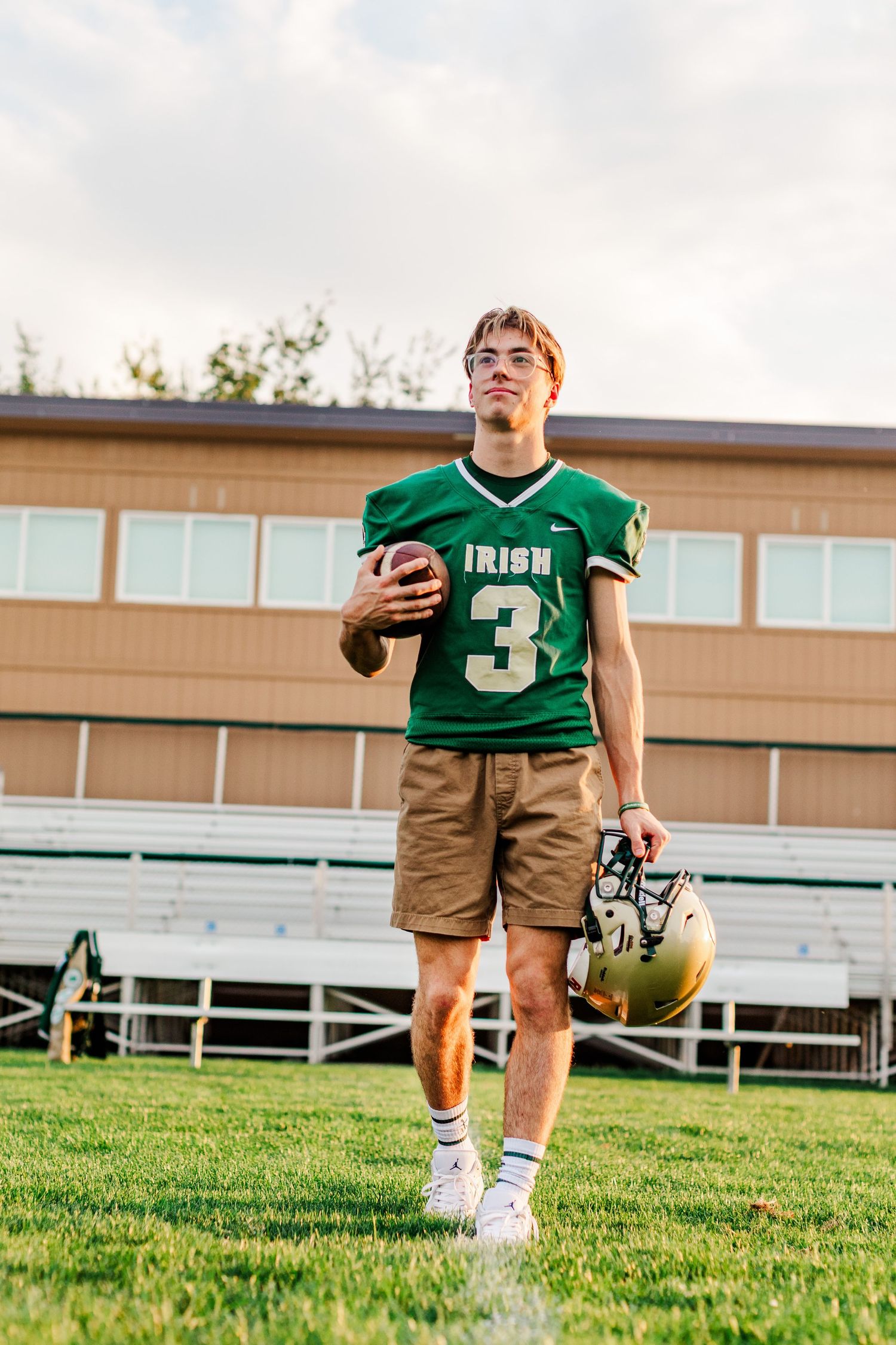 Football player in green and brown uniform stands on practice field with equipment.