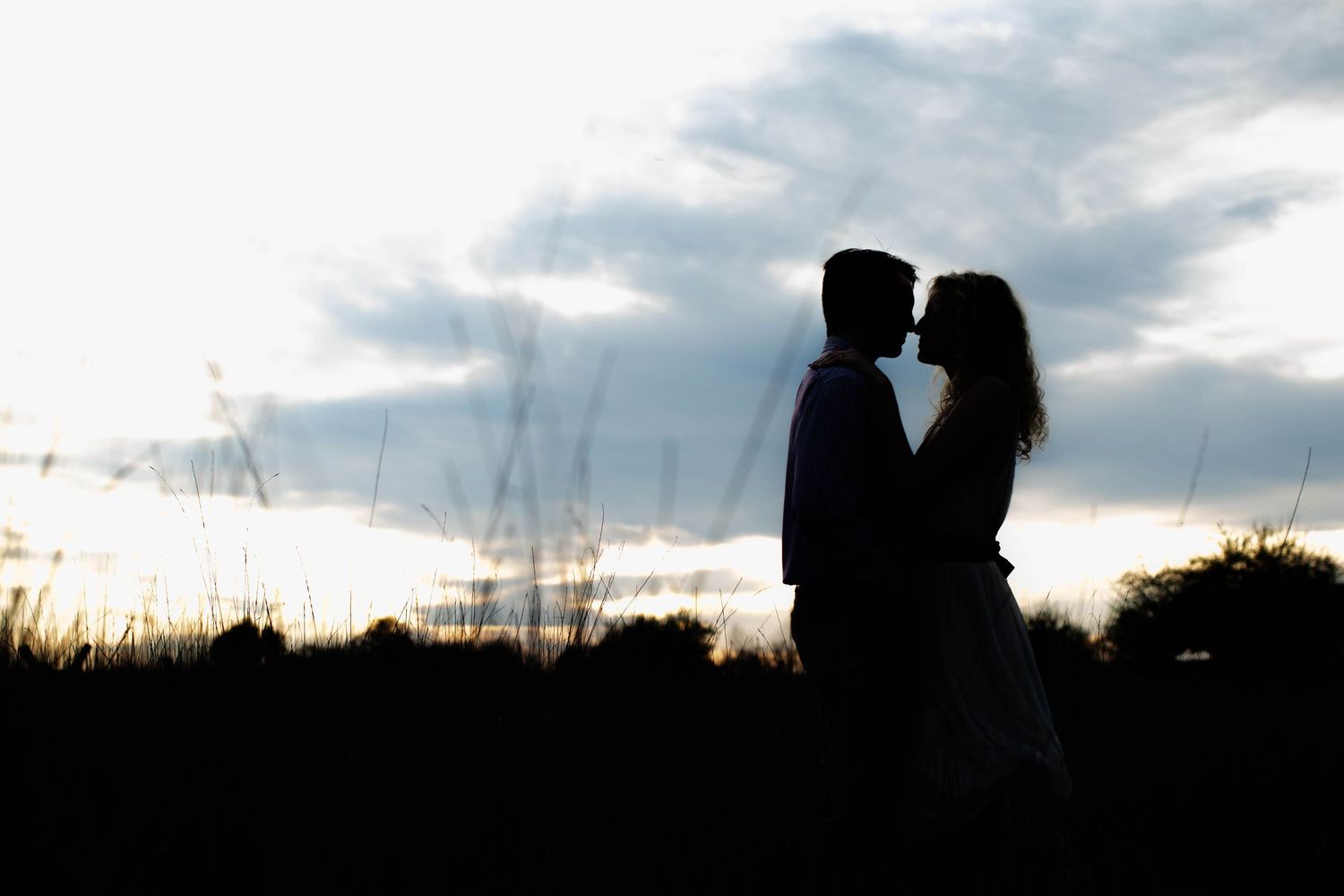 Silhouette of a romantic couple almost kissing at sunset with dramatic cloudy sky and tall grass background.