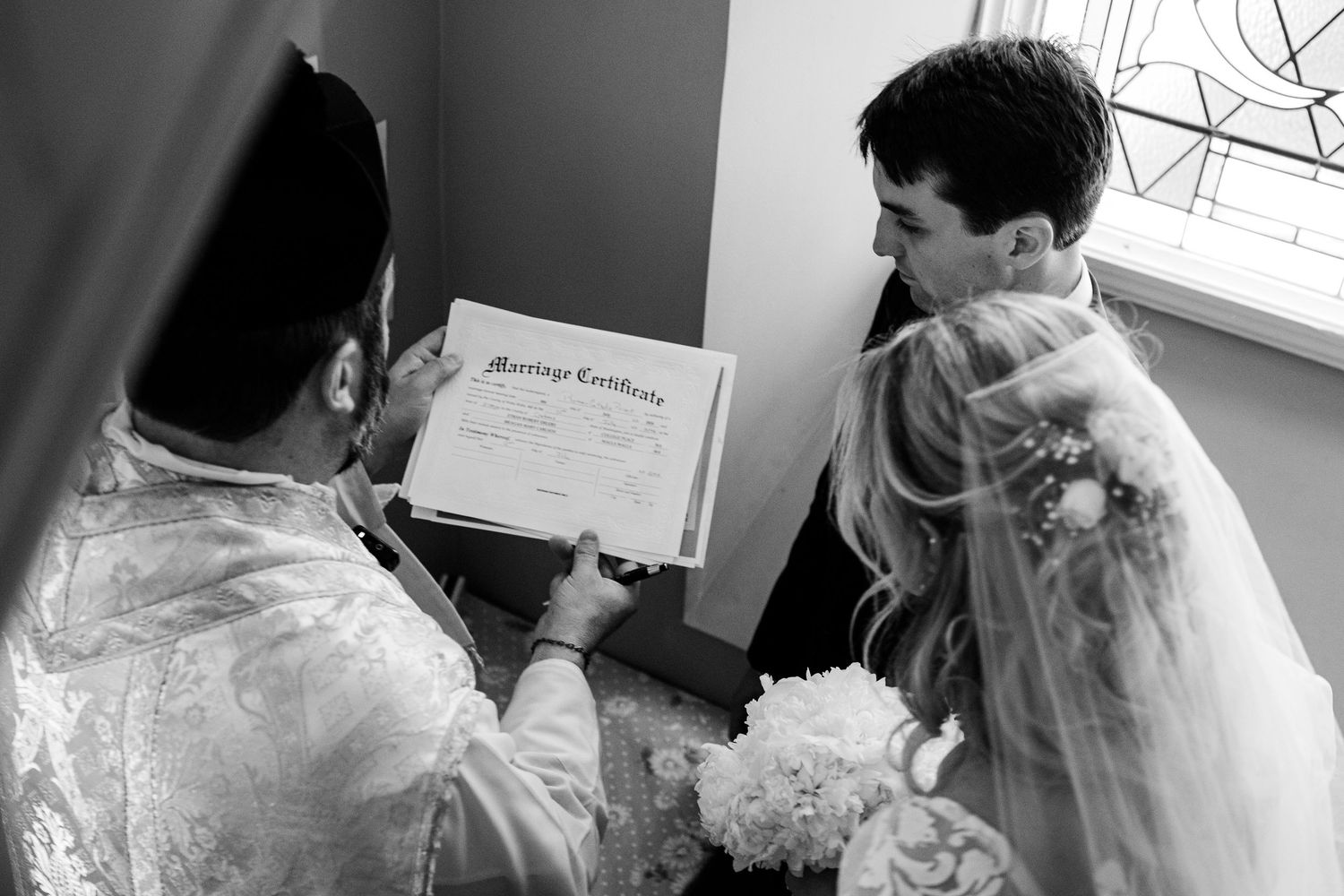Bride and groom reviewing marriage certificate with officiant during wedding ceremony in black and white photo.