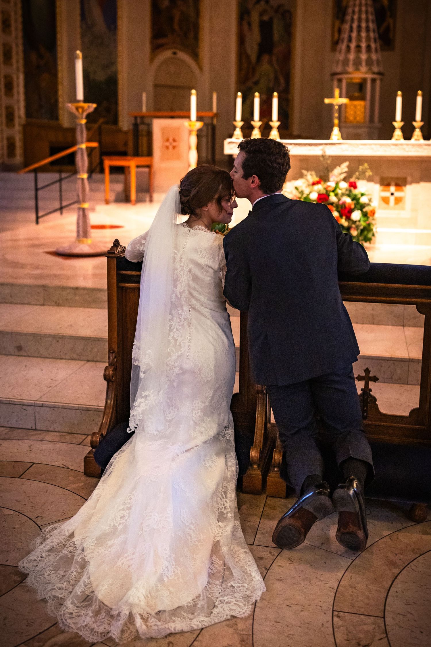 Bride and groom kneeling at church altar during wedding ceremony with glowing candles.