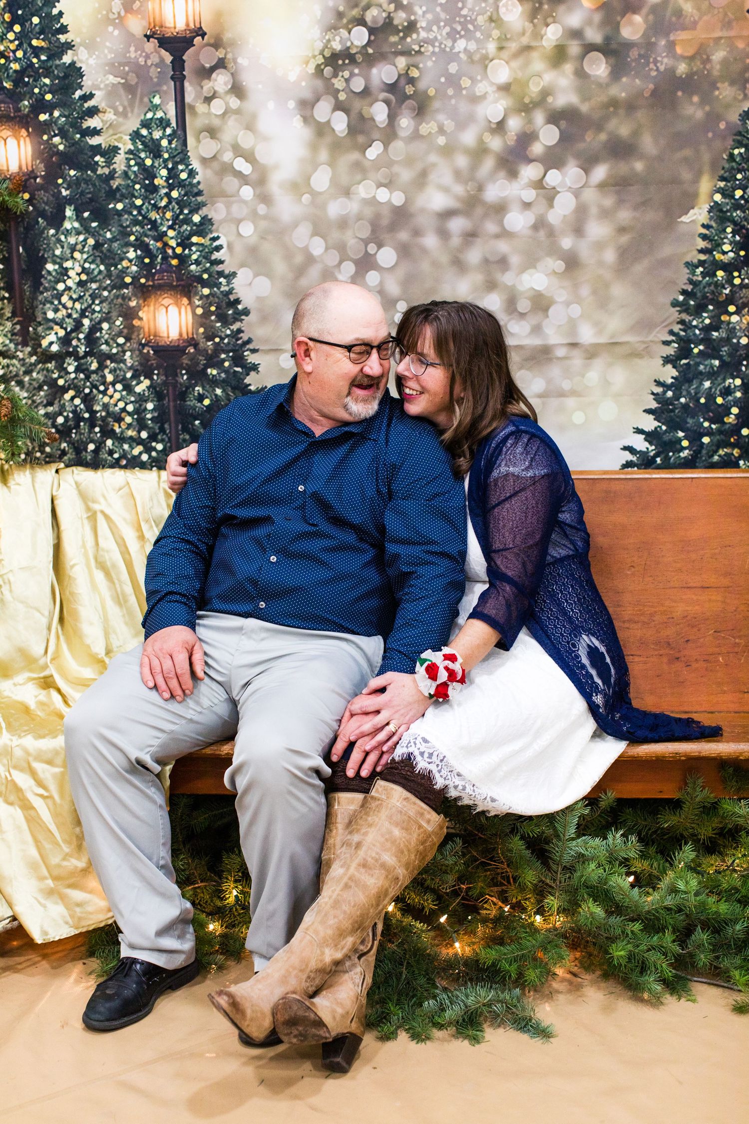 Couple shares intimate moment on wooden bench surrounded by Christmas trees and twinkling lights.
