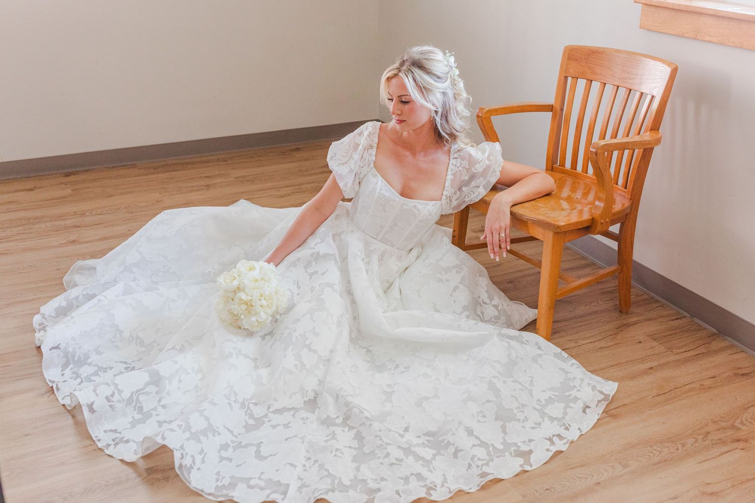 Bride in white lace wedding gown sitting on floor beside wooden chair, holding white bouquet.