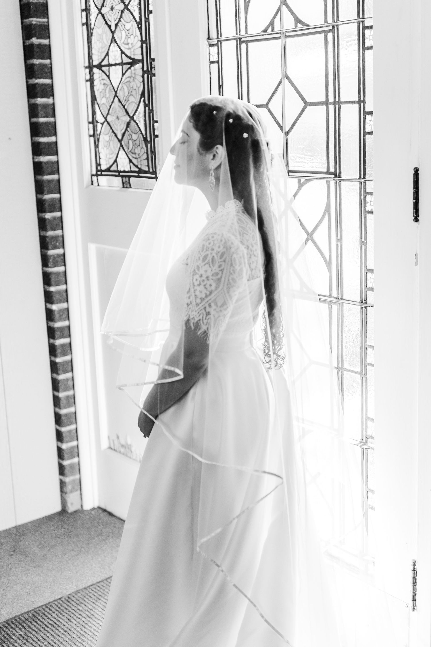 Black and white photo of a bride in a lace wedding dress and veil standing by a decorative window.