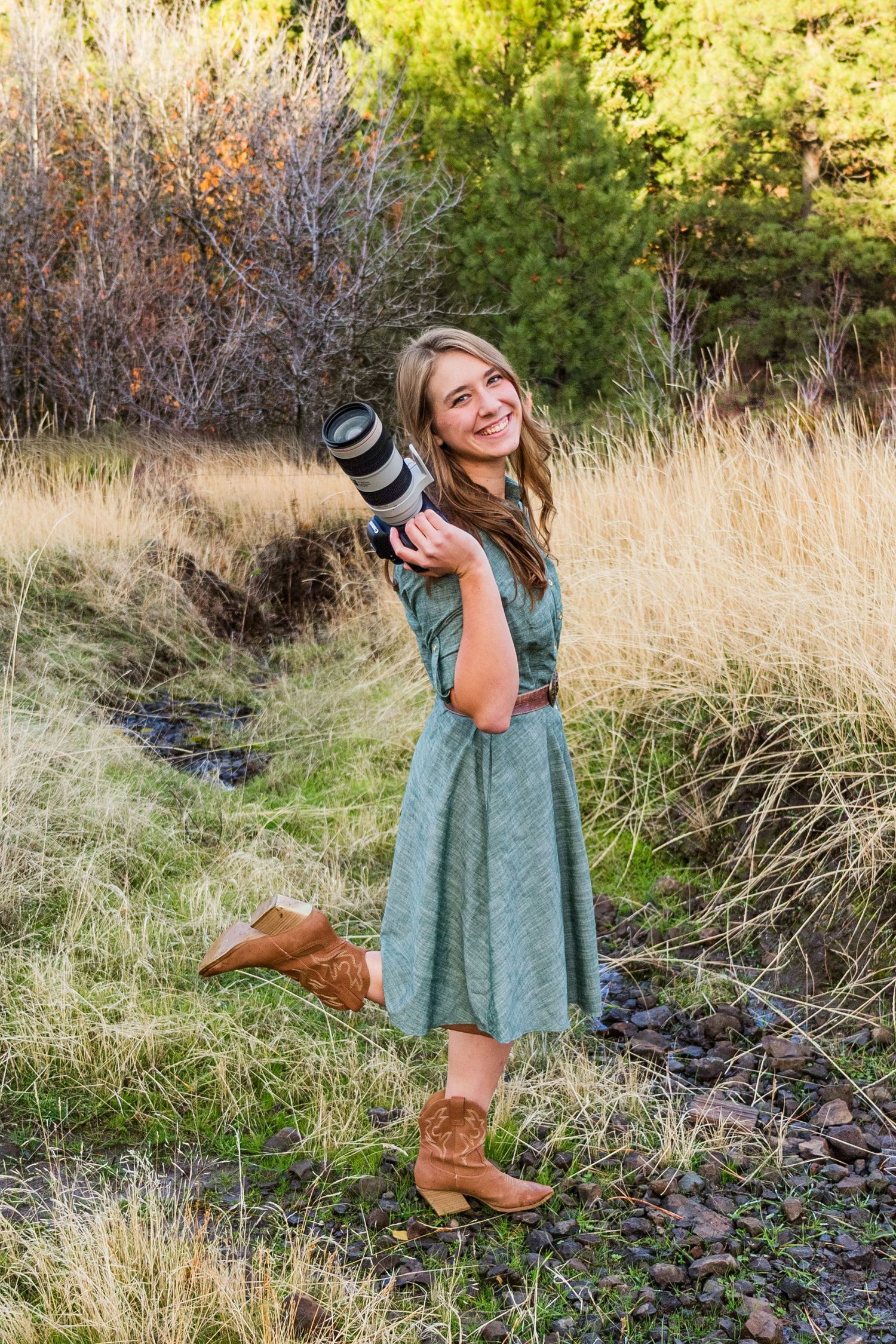 A photographer in a green dress and brown boots poses playfully in a golden autumn field with camera gear.