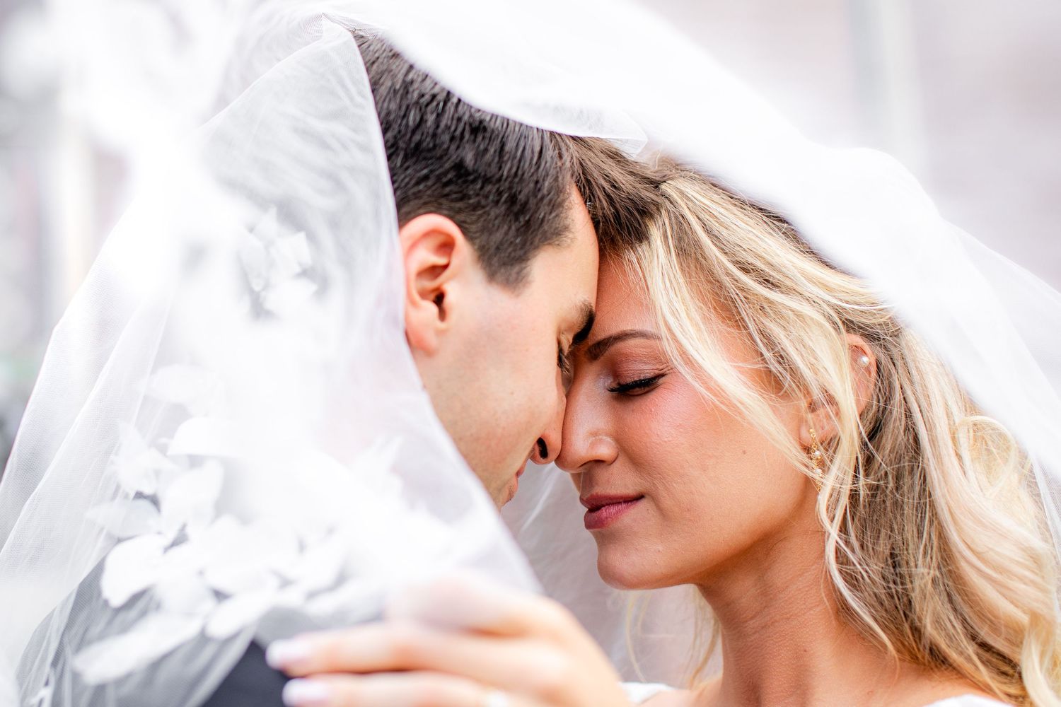 Bride and groom share an intimate moment under a white wedding veil, foreheads touching tenderly.