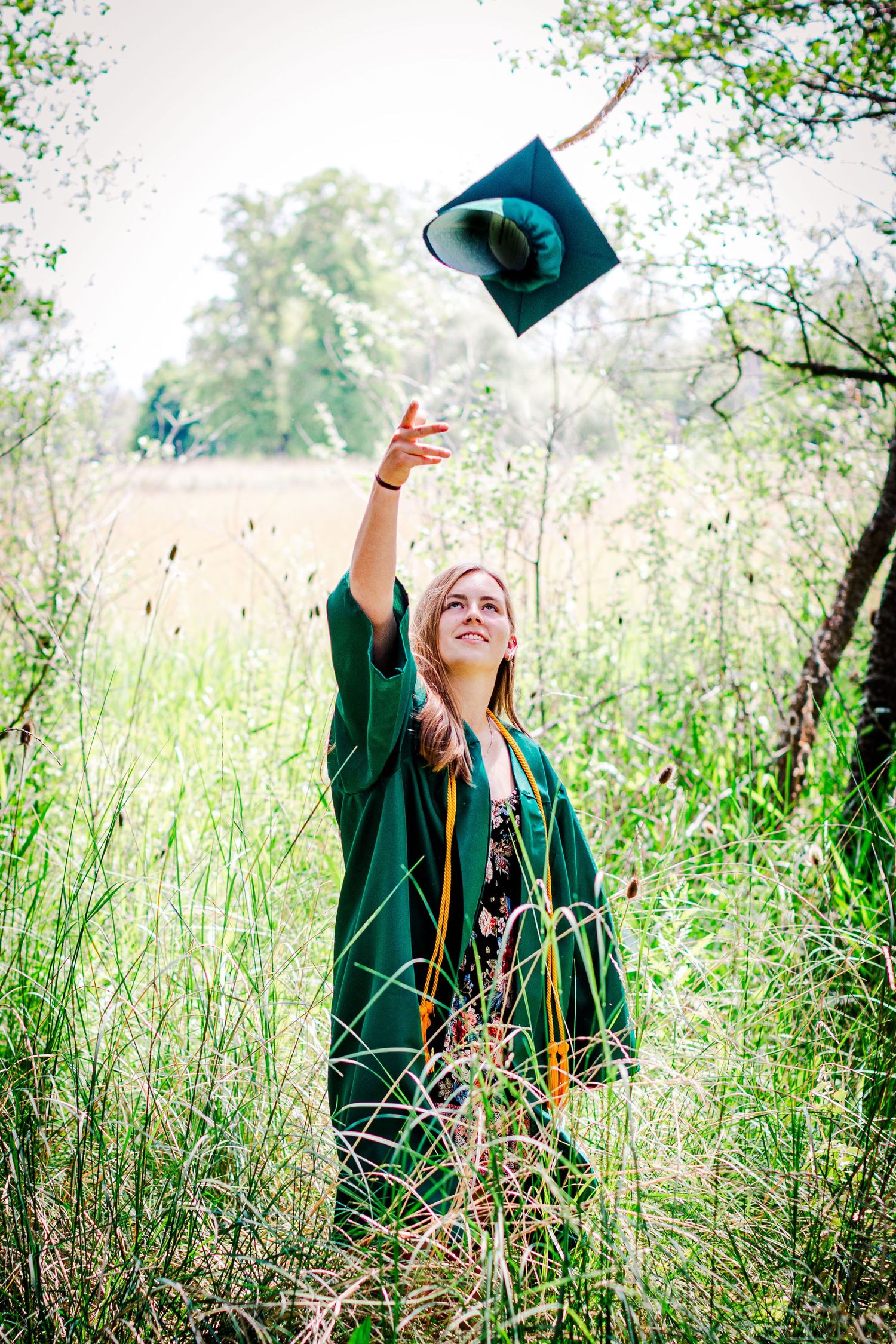 A DeSales Catholic High School graduate in emerald green regalia celebrates in a sunlit meadow with tall grass.