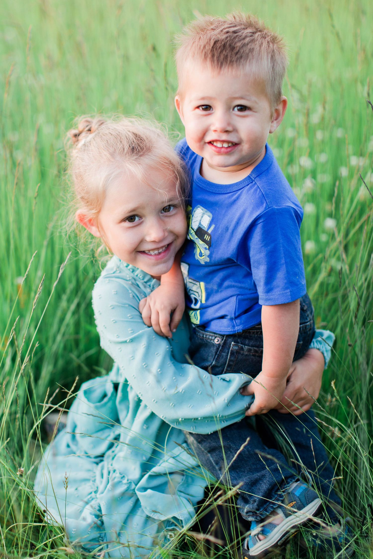 Two young children playing together in tall grass wearing blue and turquoise clothing.