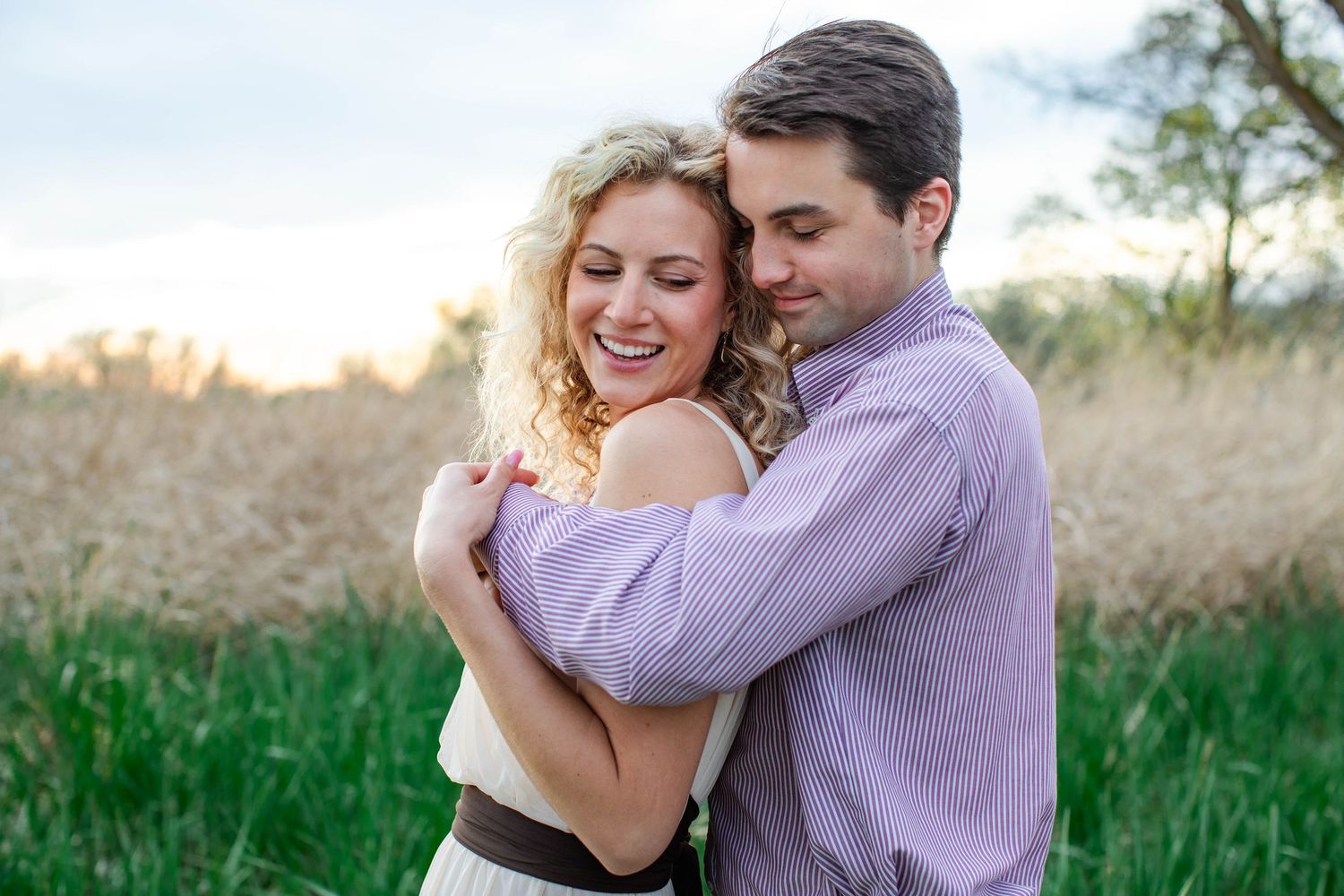 A smiling couple embraces warmly in a field at sunset, the woman in a white dress and man in a purple shirt.