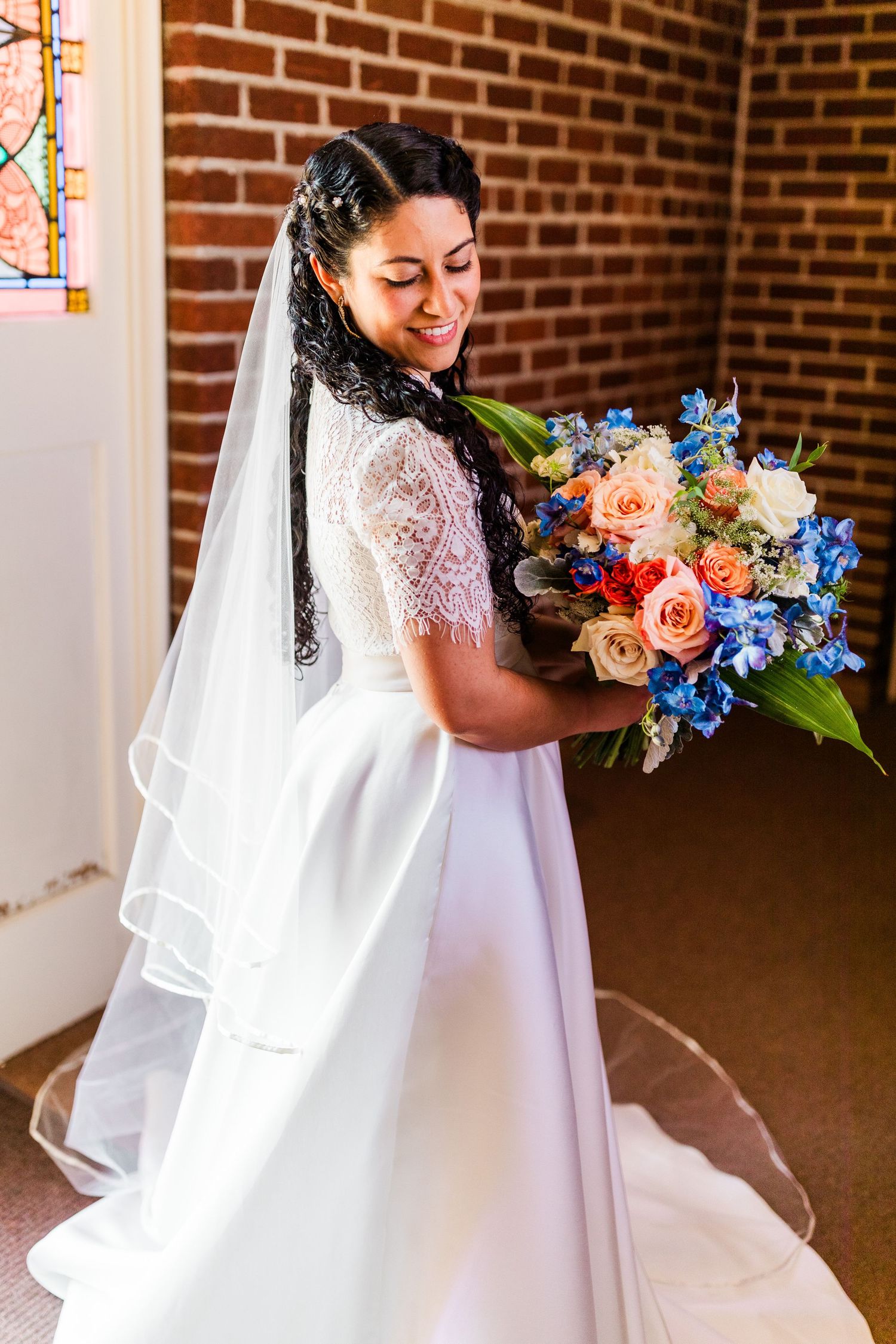 Bride in a lace wedding dress holding a vibrant bouquet with blue and peach flowers against a brick wall.