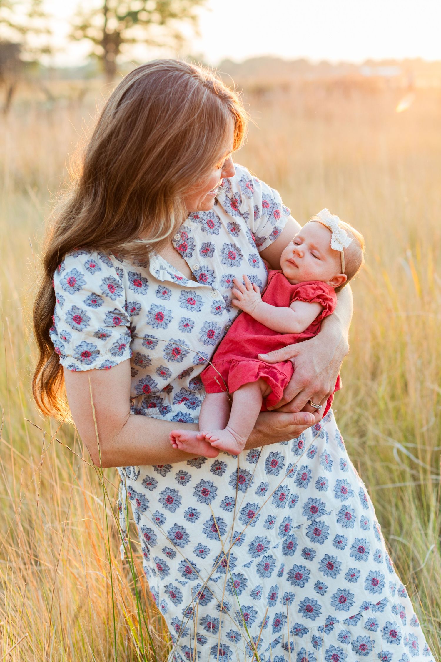 Mother in floral dress holding newborn baby in red outfit in golden sunlit field.