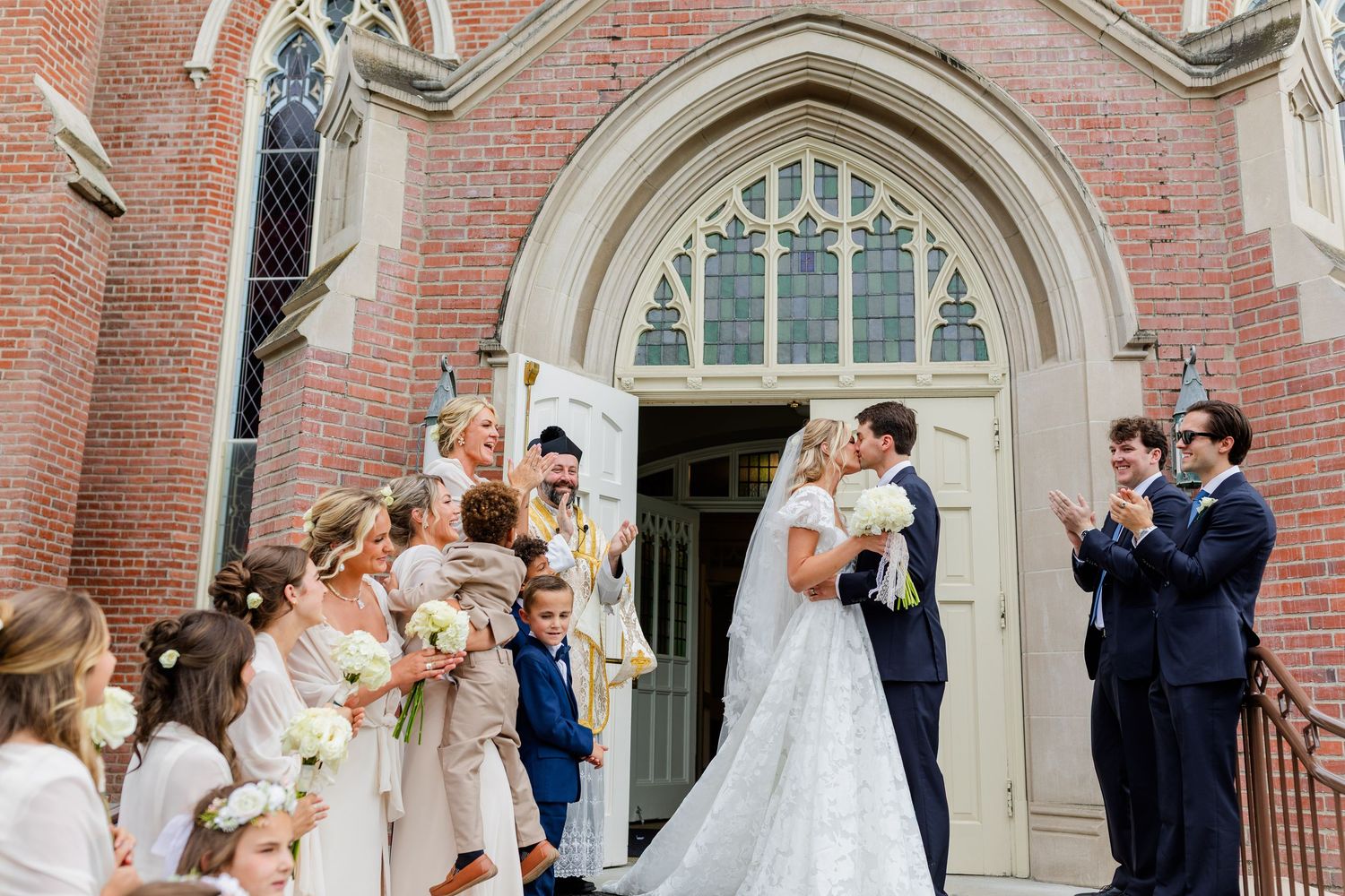 Bride and groom share a kiss outside a brick church surrounded by bridesmaids in champagne gowns and groomsmen in navy suits.