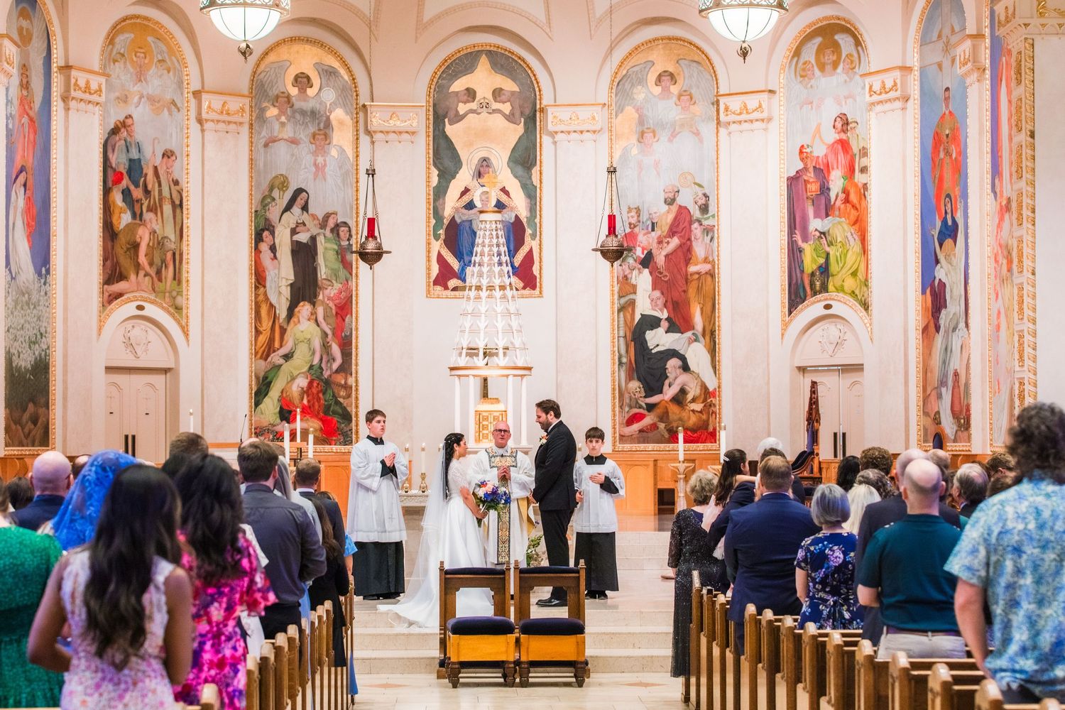 Church wedding ceremony with ornate religious murals and gathered guests in colorful attire.