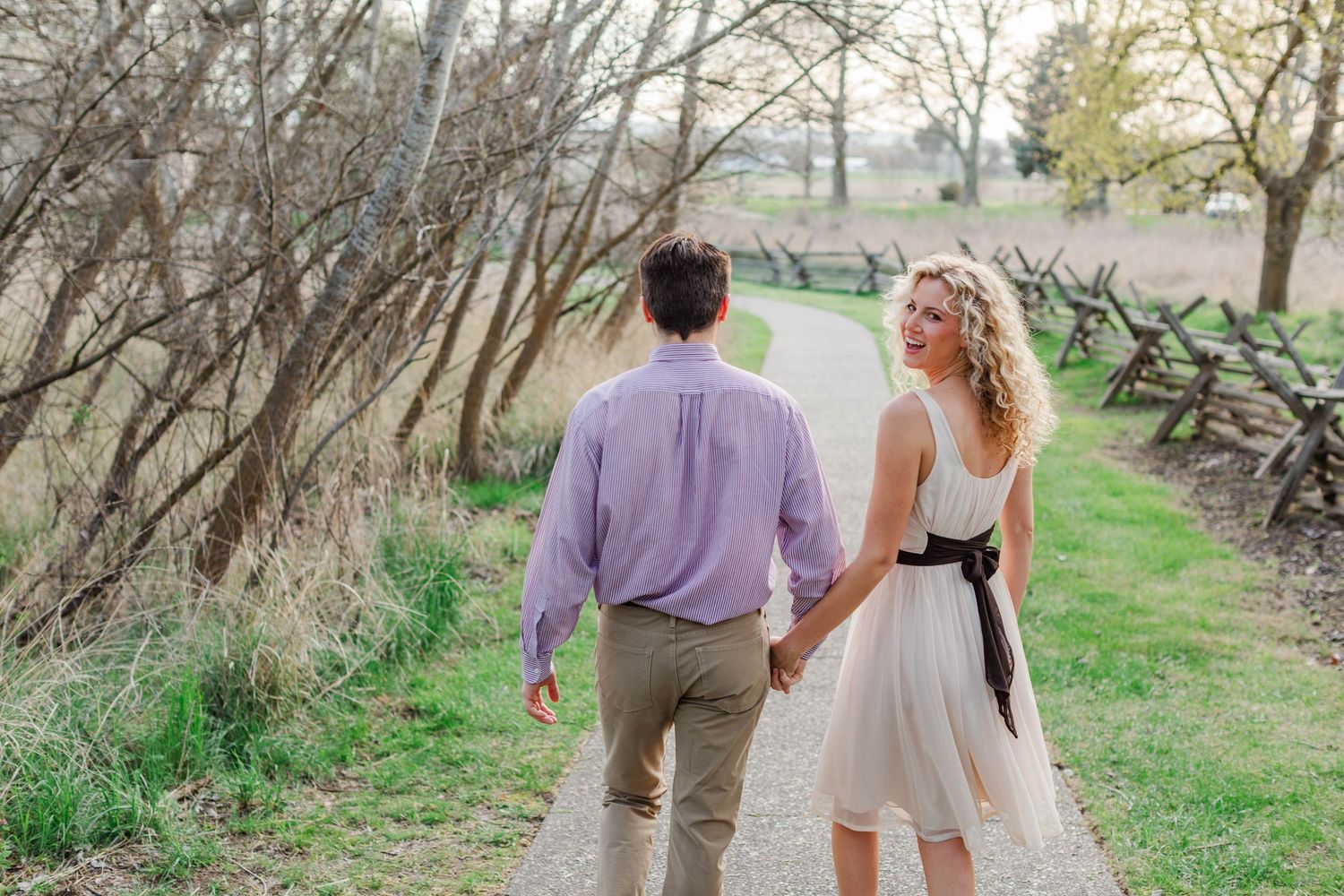 A couple walks hand-in-hand on a scenic path, the woman in a white dress glancing back, surrounded by lush greenery.